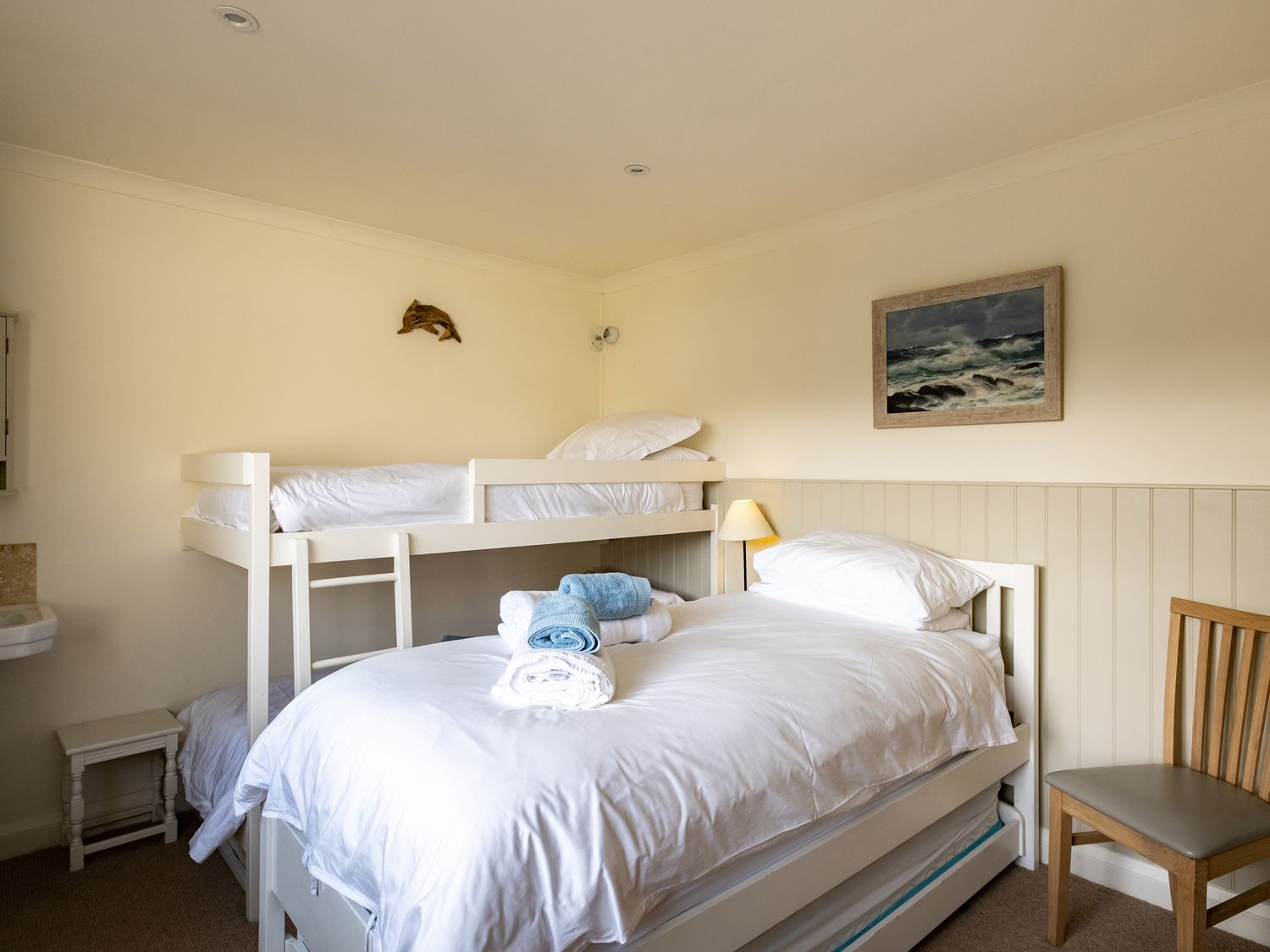 A bedroom with a bunk bed and a single bed with folded towels a wooden chair a sink and wall art at Atlantis in Polzeath