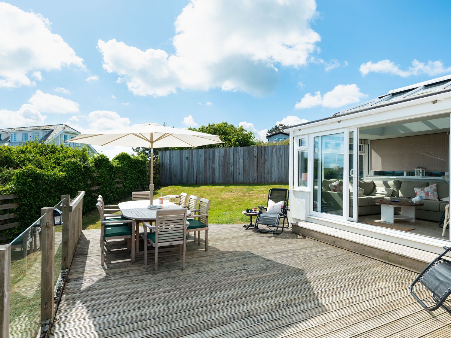 A wooden deck with a dining table and chairs under a large umbrella next to a garden and a room with a sofa at Atlantis in Polzeath