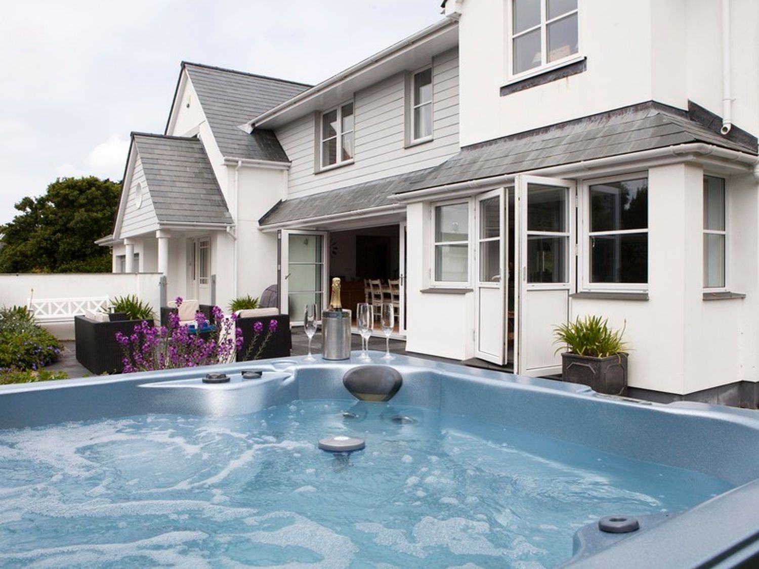 An outdoor hot tub with a bottle of champagne and glasses next to a white house with open doors and garden furniture with flowers at Cherrytrees in Rock