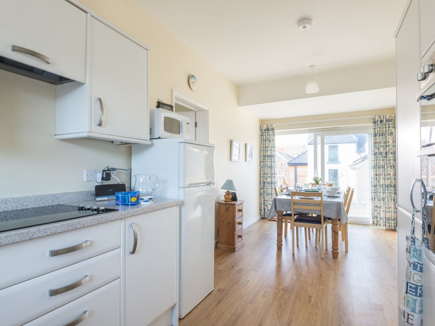 A kitchen area with white cabinets a dining table set with chairs and large window at The Holiday House (2 Atlantic Mews) in Polzeath