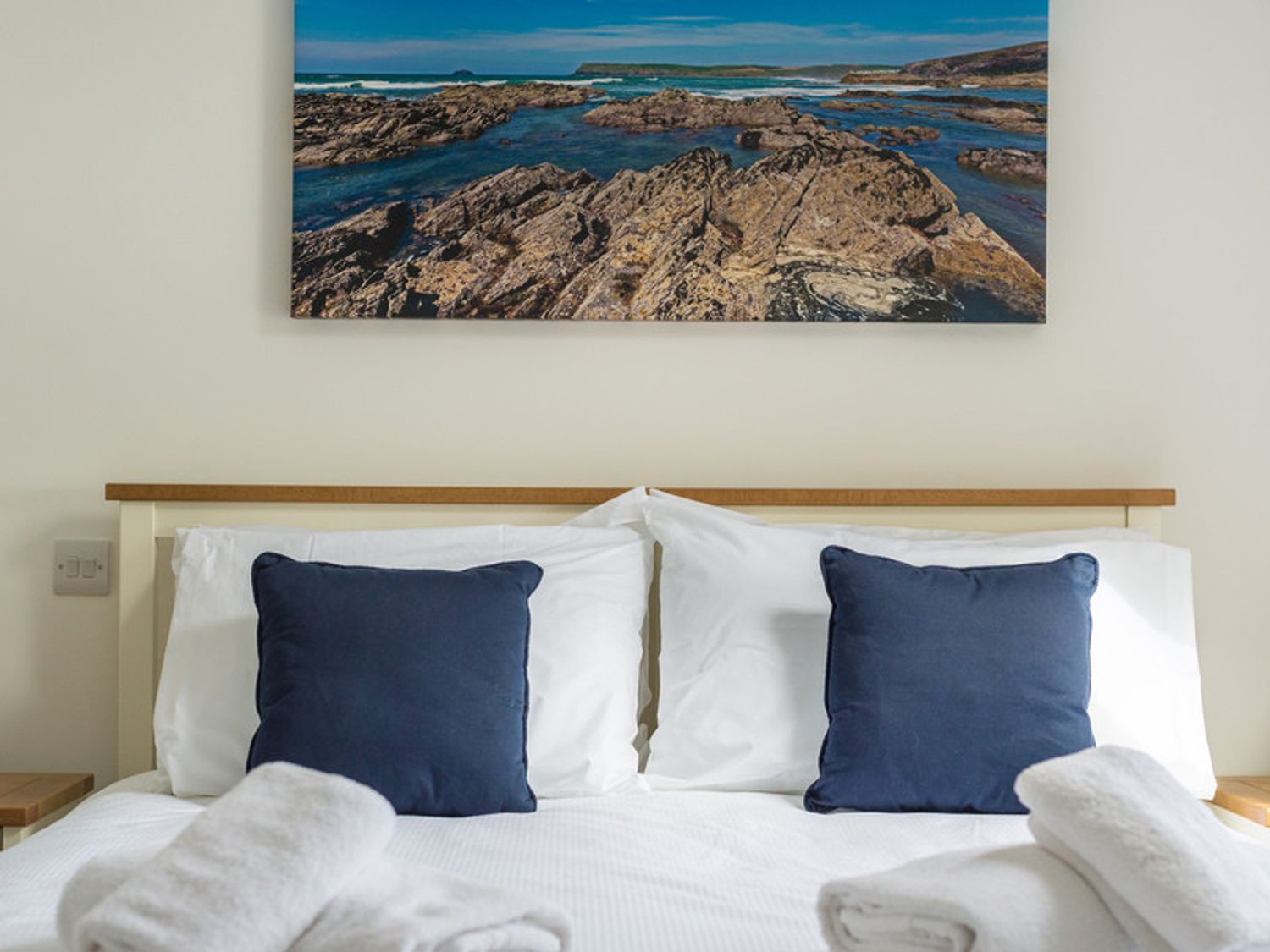 A bed with white sheets blue pillows and folded white towels under a wall photo of rocky coastline at Rainer Rocks in Polzeath