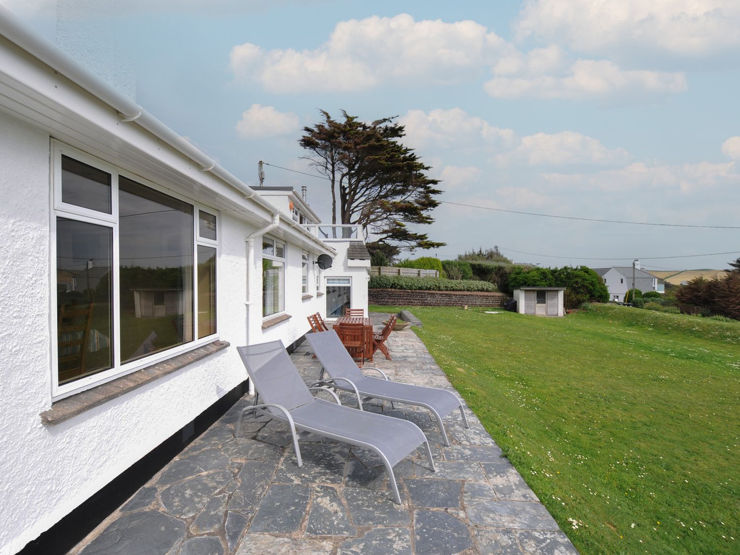 A patio with two lounge chairs and a wooden table set beside a white house with large windows and a grassy lawn at Dolphins in Daymer Bay