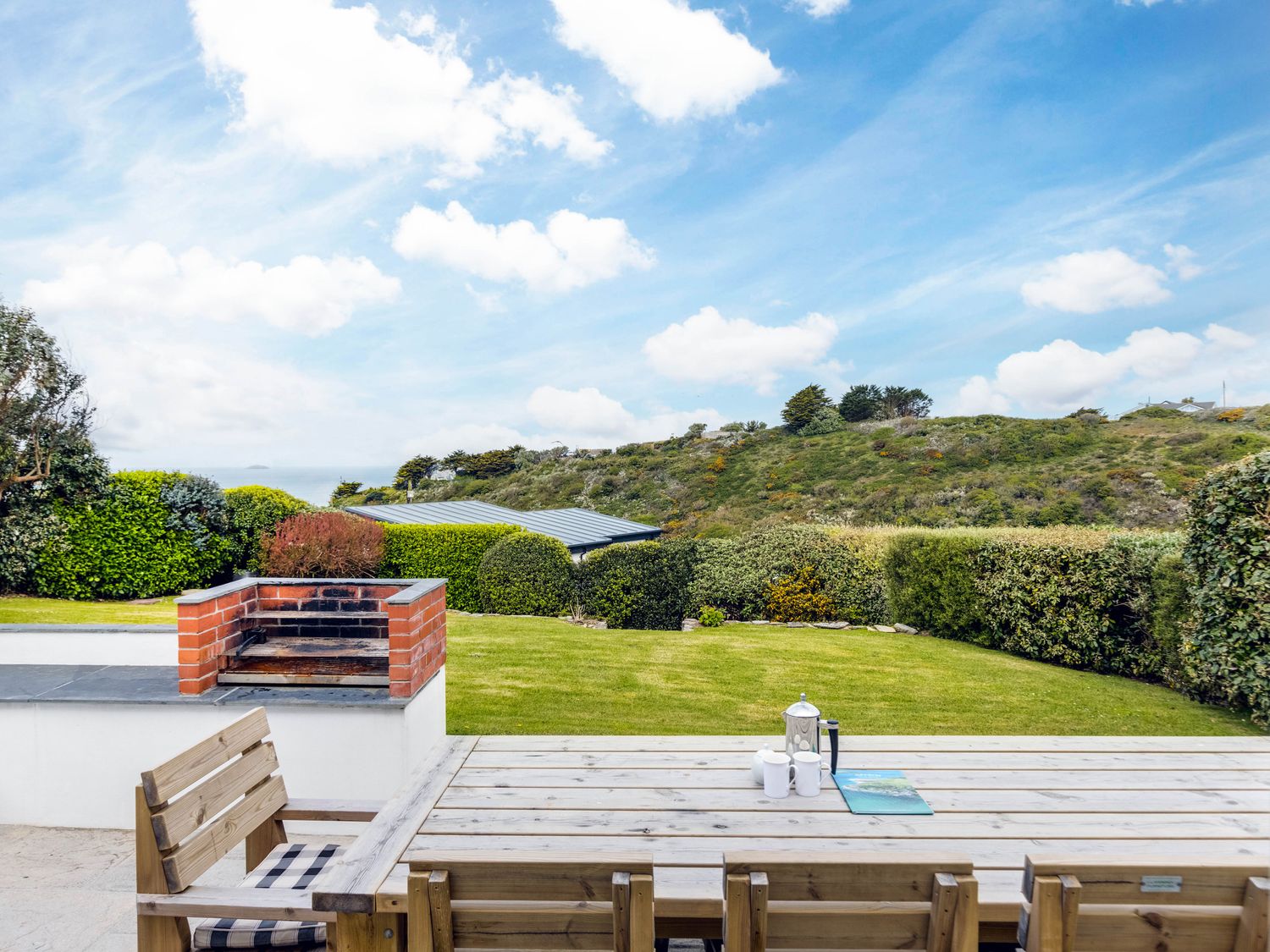 An outdoor patio with wooden table and chairs a built in brick barbecue and green garden with bushes and a hill in the background at Westpoint in Polzeath