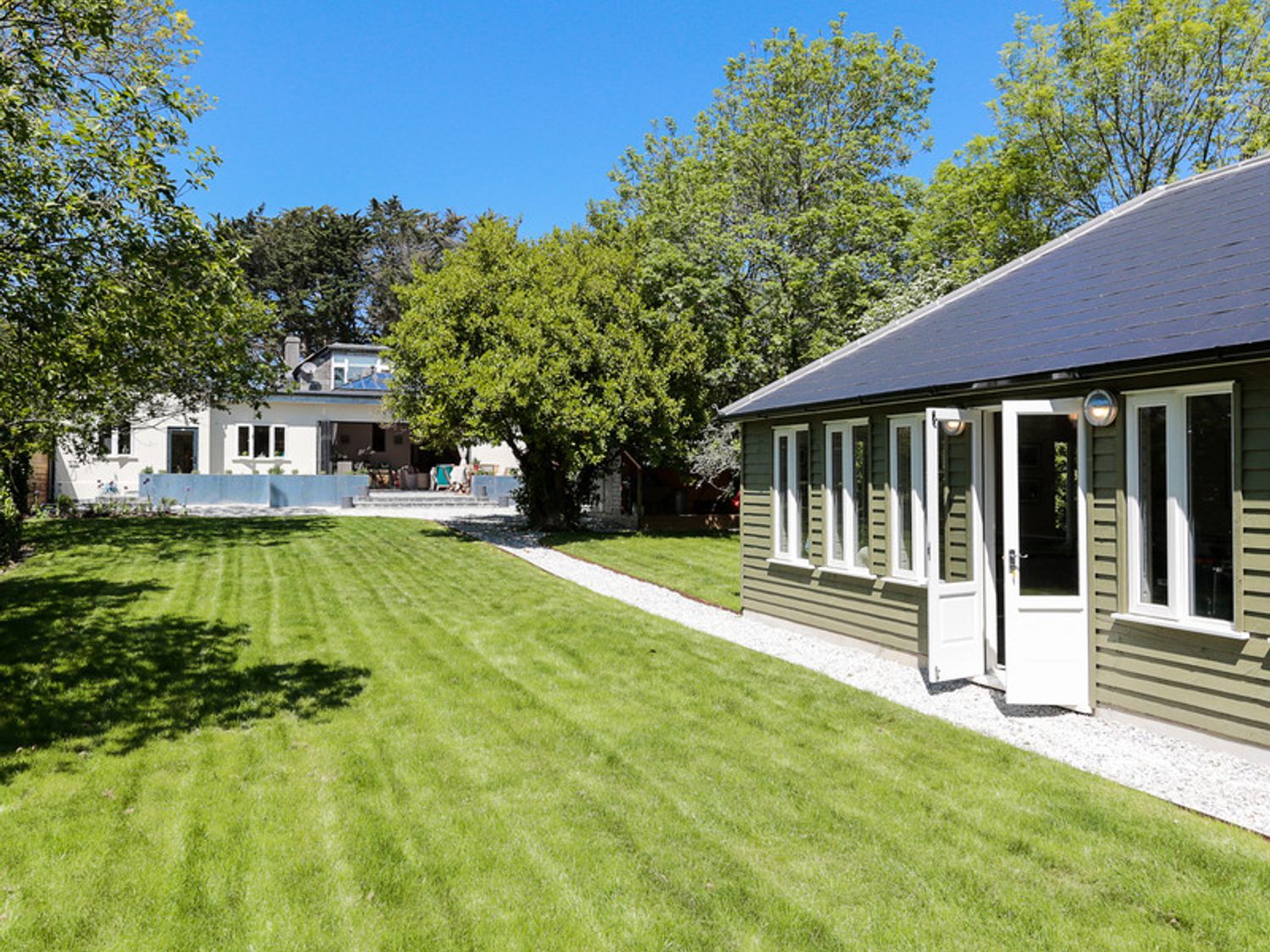 A green lawn with a pathway between a white house and a green building with multiple windows at Trevanion in Rock
