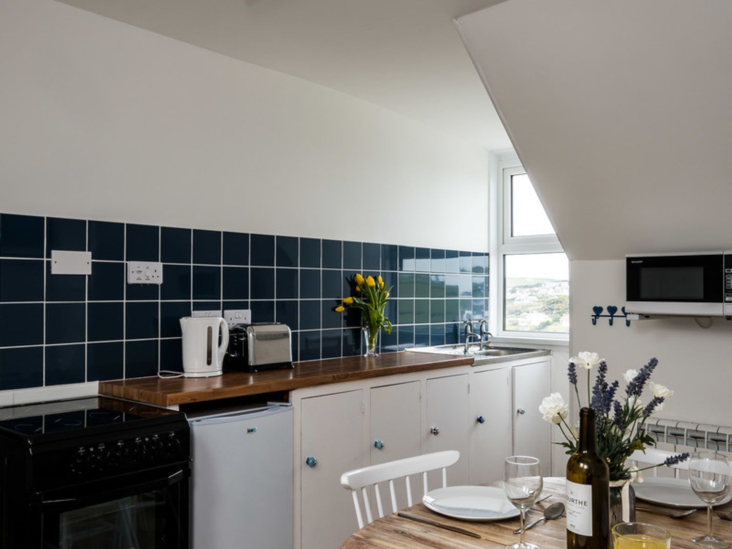 A kitchen with blue tiled backsplash a kettle toaster flowers on the counter a microwave and a dining table with plates glasses and flowers at Pinewood 14 in Polzeath