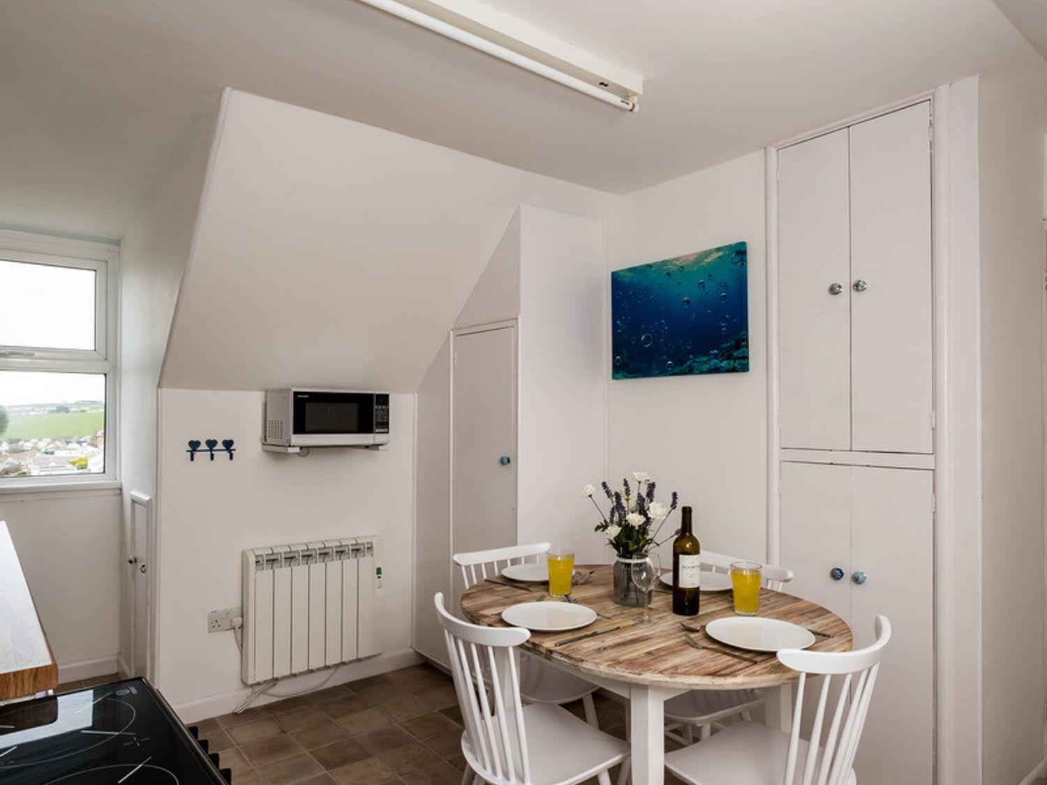 A kitchen dining area with a round table set for four, white chairs, a microwave on the wall, cupboards, and a window at Pinewood 14 in Polzeath