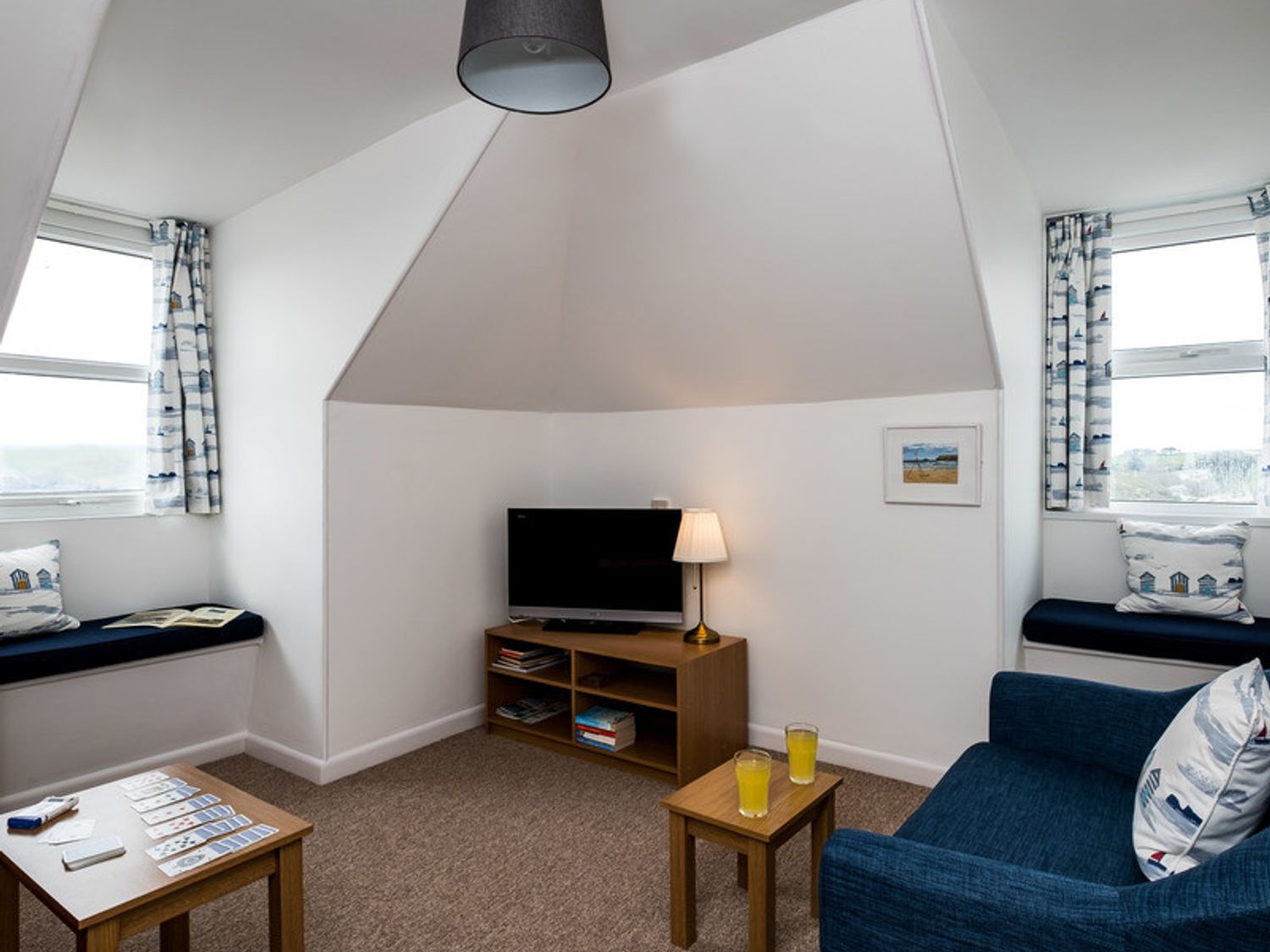 A small sitting room with a television on a wooden stand cushions on window seats and two small tables with drinks at Pinewood 14 in Polzeath