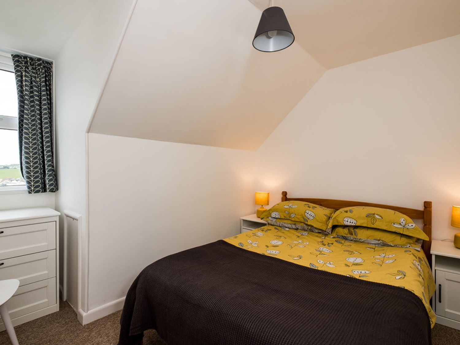 A bedroom with a bed covered with yellow floral bedding two bedside tables with lamps a chest of drawers and a window with patterned curtains at Pinewood 14 Polzeath