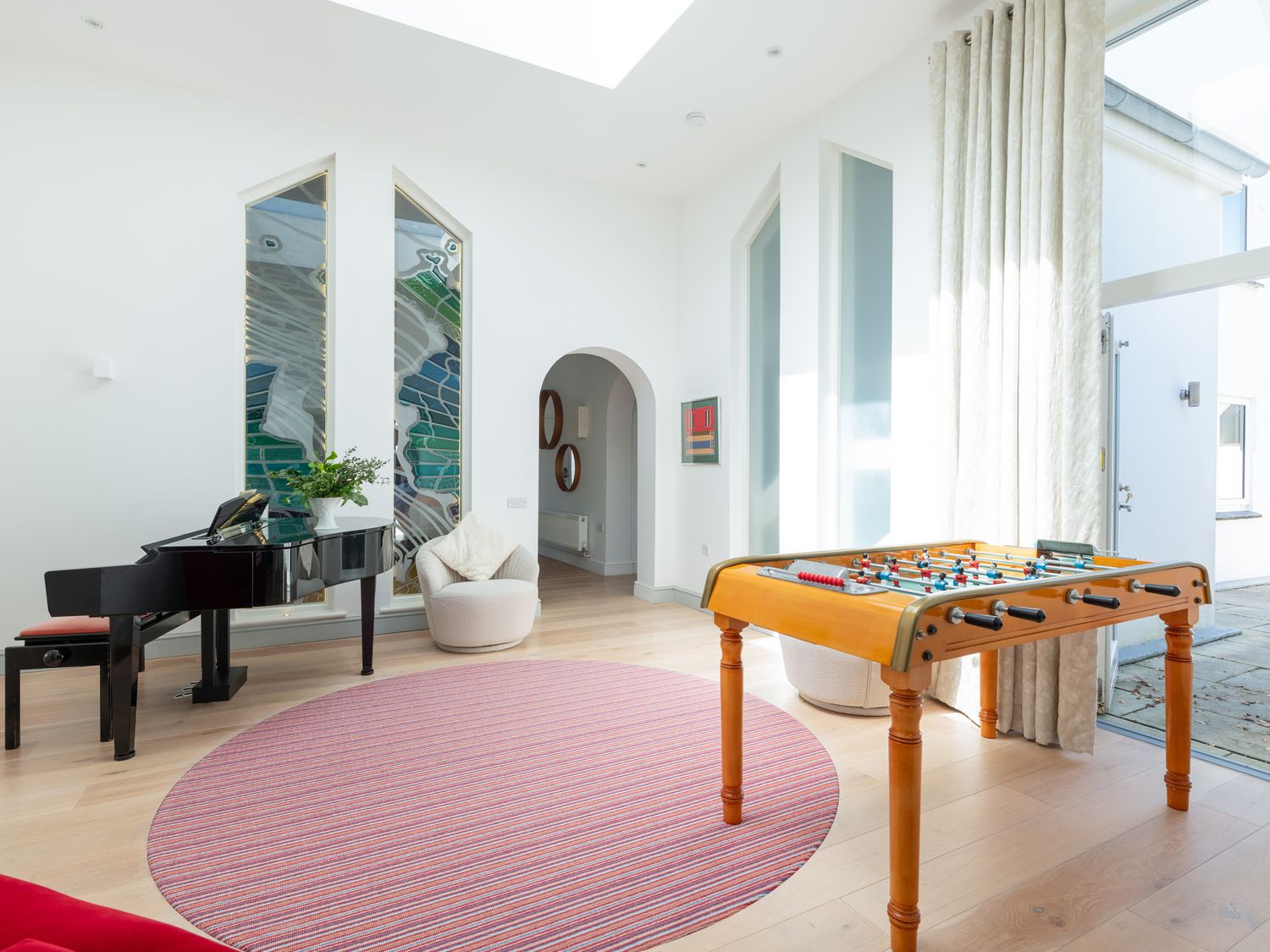A room with a black grand piano, a wooden foosball table, a round pink rug, and a white chair near arched doorway at Landers Field in Rock