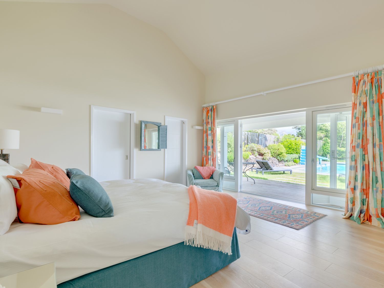 A bedroom with a bed with orange and green pillows a green chair near glass doors leading to a garden and pool with patterned curtains at Landers Field in Rock