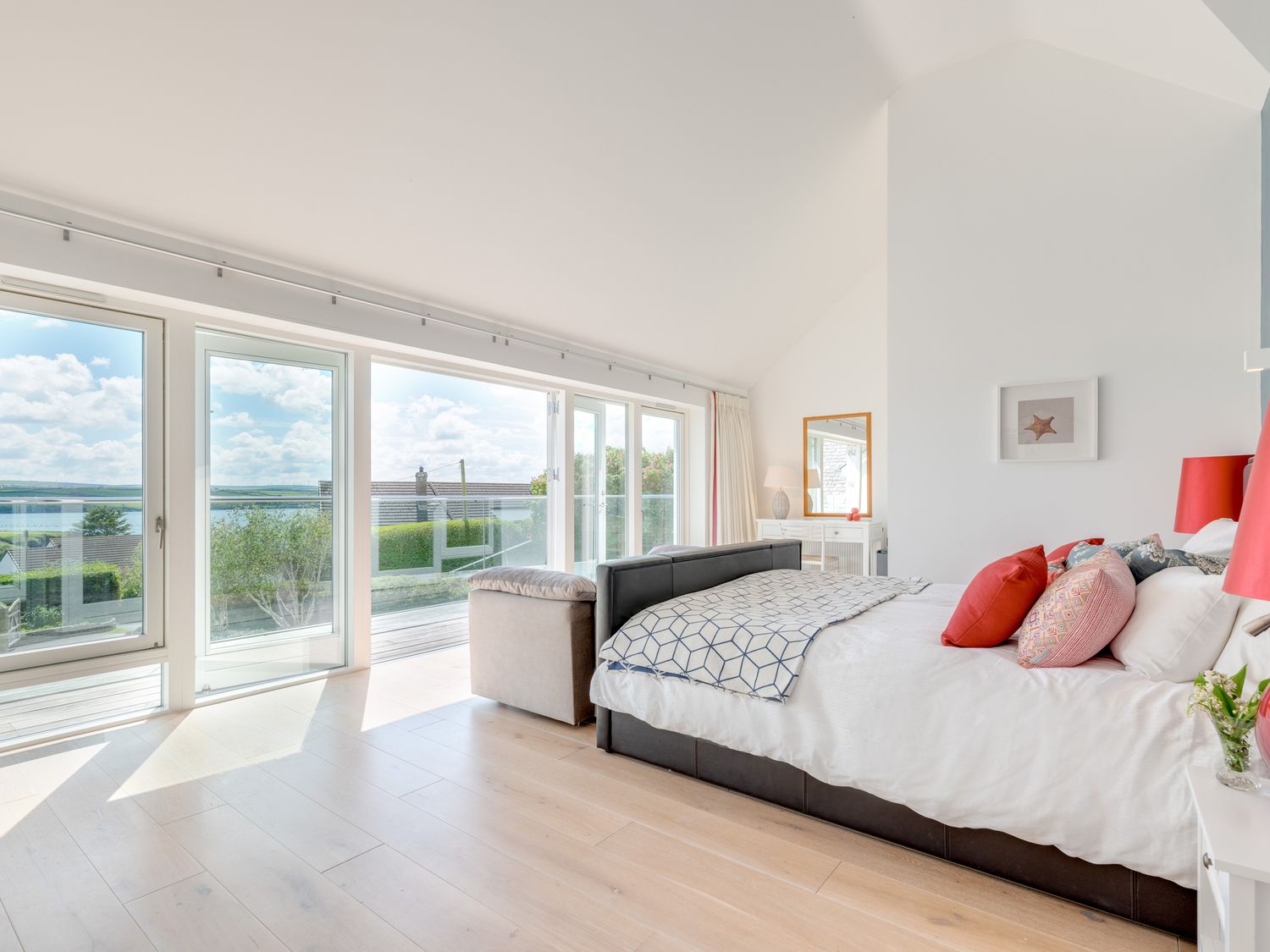 A bedroom with a bed and red cushions next to large windows overlooking a garden and water at Landers Field in Rock