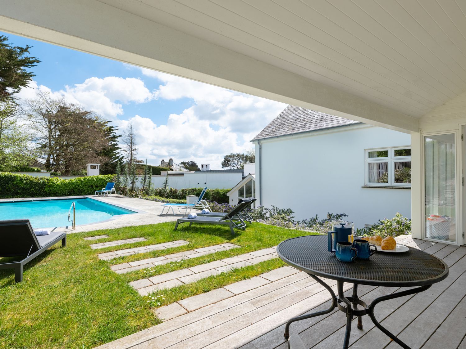 A covered patio with a round table set with cups and croissants overlooking a grassy yard with lounge chairs and a swimming pool at Landers Field in Rock