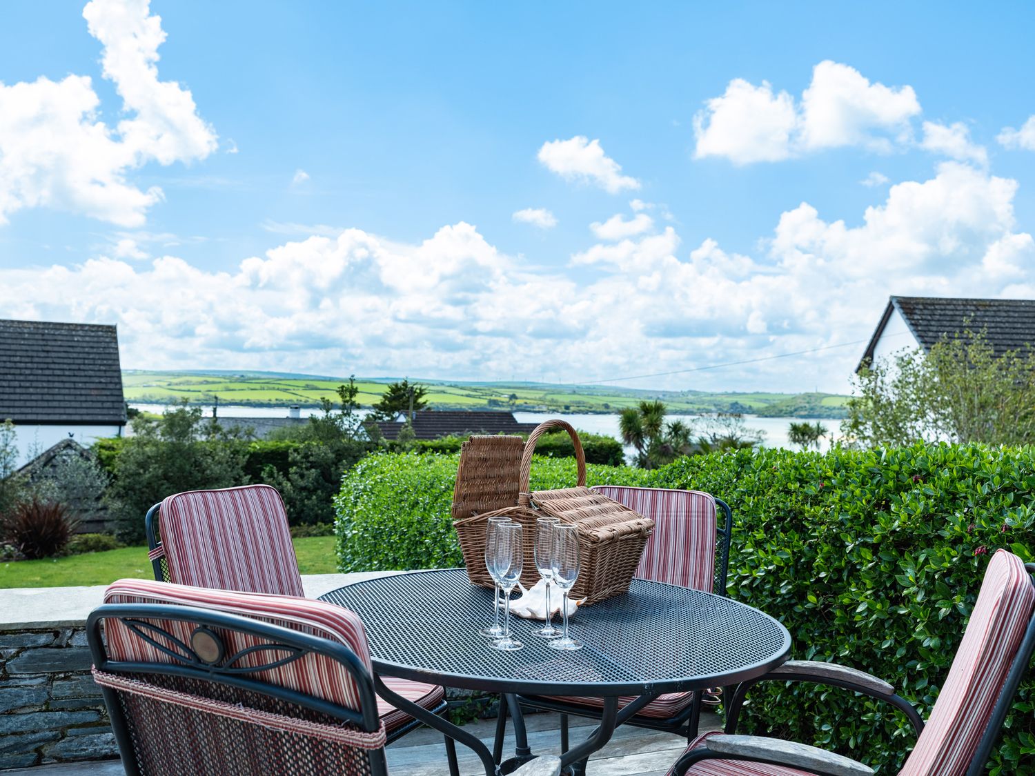 An outdoor patio with a round metal table, four striped cushioned chairs, a wicker picnic basket, and four empty glasses at Landers Field in Rock