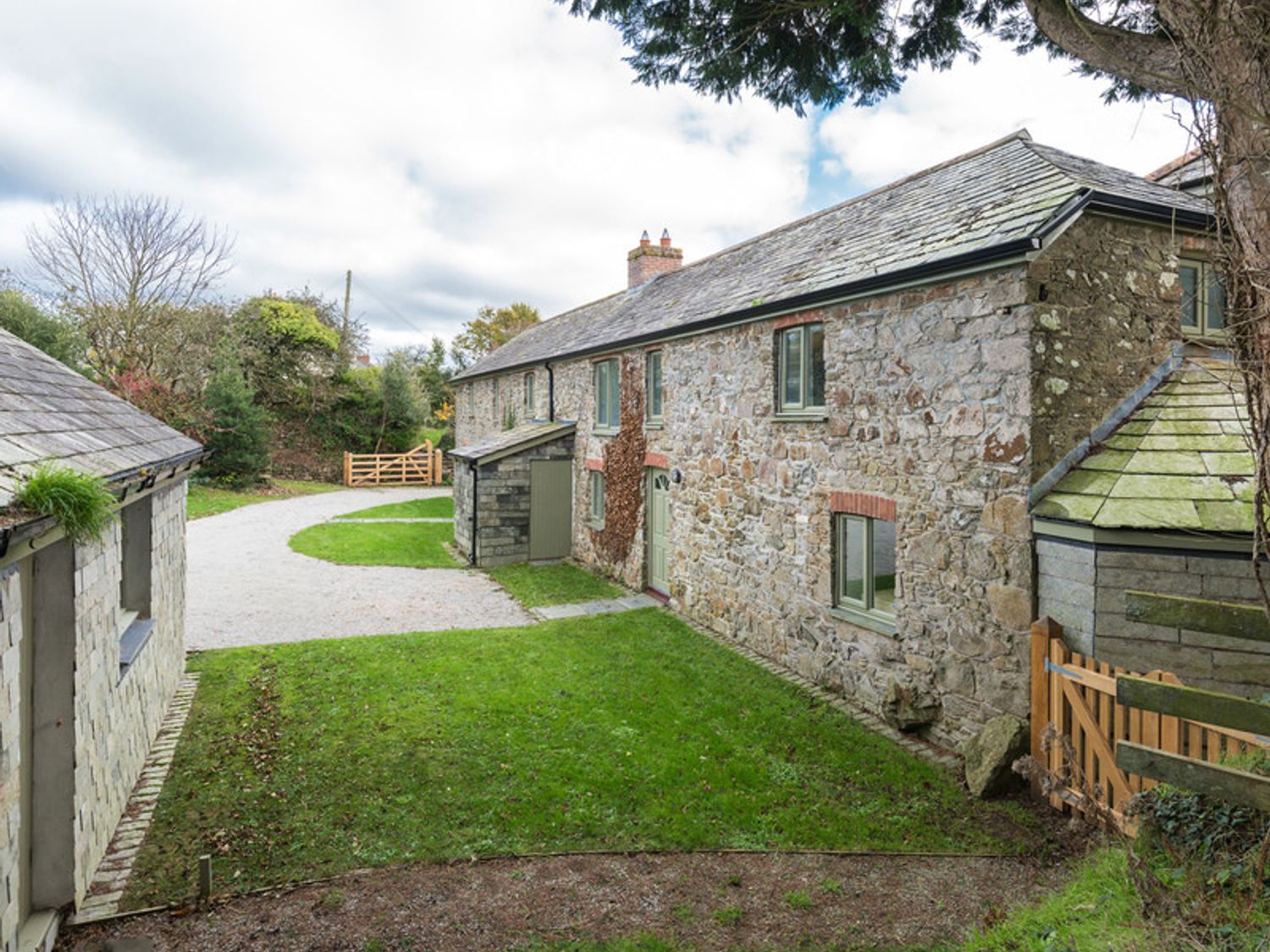 An outdoor view of a stone cottage with grass lawn gravel driveway and wooden gates at Orchard Cottage in Polzeath