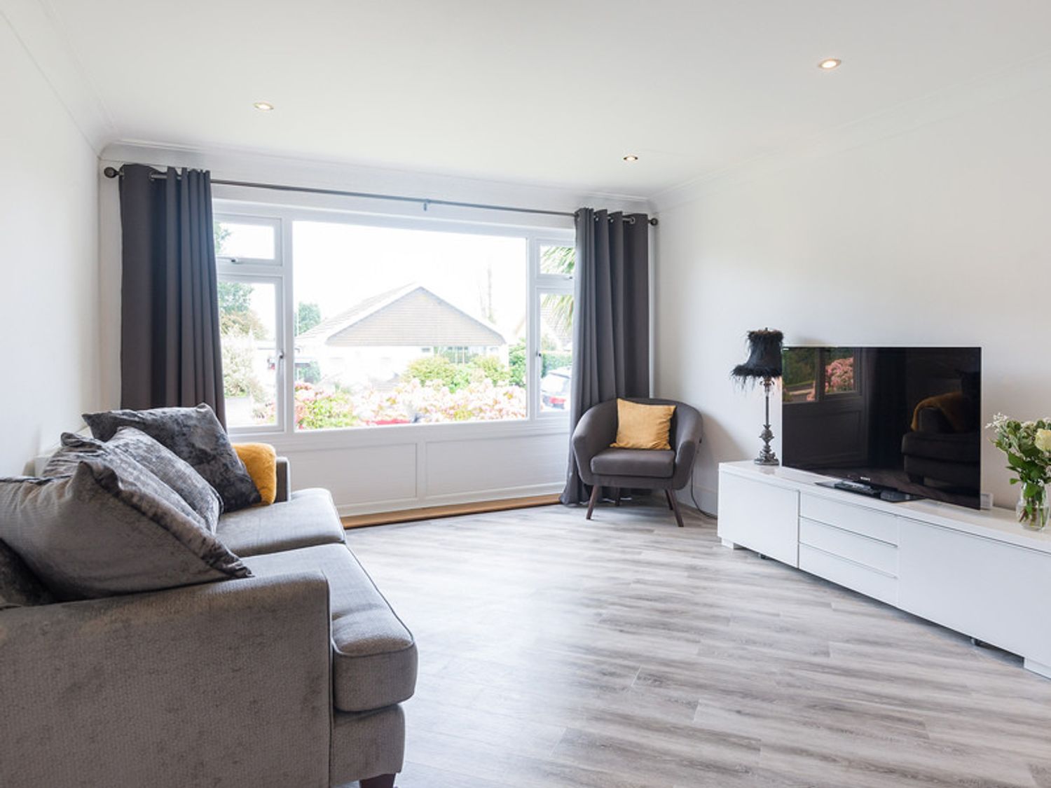 A living room with a gray sofa and pillows a gray armchair with a yellow cushion a large window with dark curtains a white TV stand a flat screen TV and a small lamp with a black shade at Greenaway 4 Rockhaven Gardens Rock