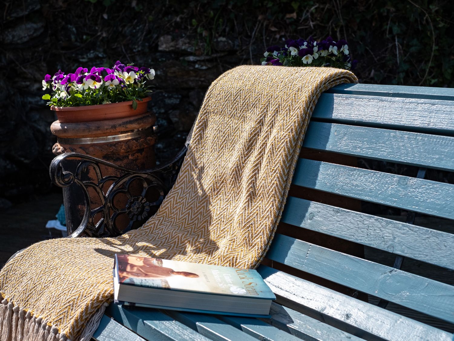 A blue wooden bench with a yellow patterned blanket and a book with a flower pot on an iron stand behind the bench at Grey Roofs in Port Isaac