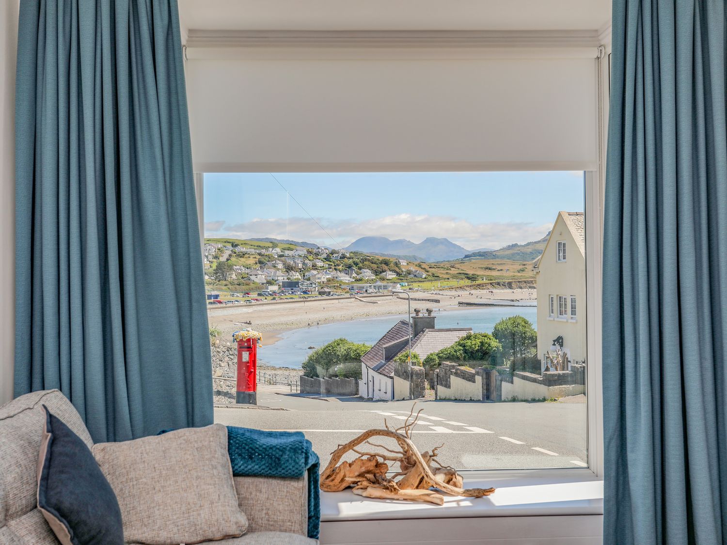 A window view of a coastal town with a red pillar box and houses from a living room with blue curtains and a beige sofa at Gwynle - Criccieth in Criccieth