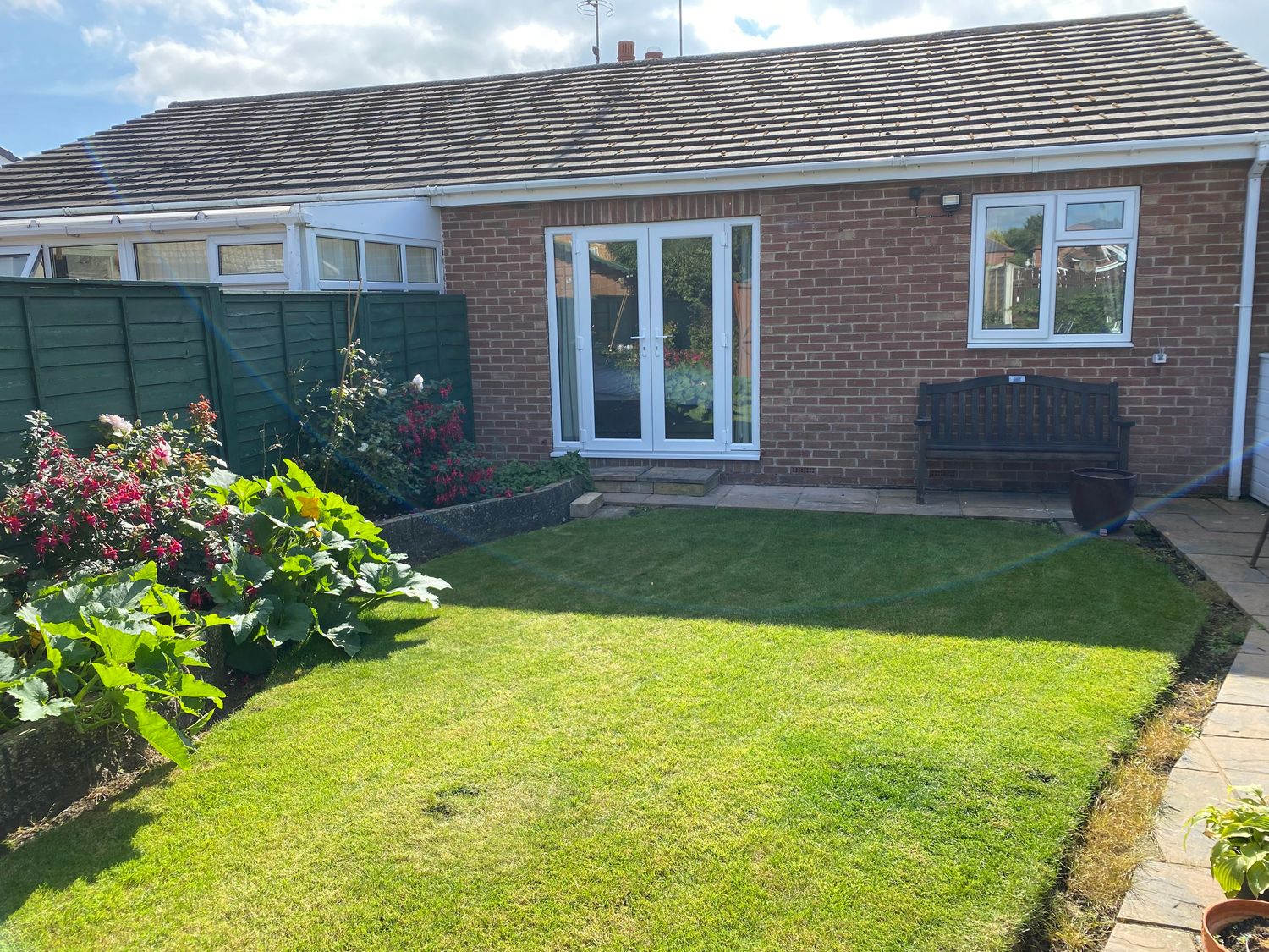 A backyard with a lawn flowers a wooden bench and a brick house with glass doors at The Corner Bungalow in Driffield