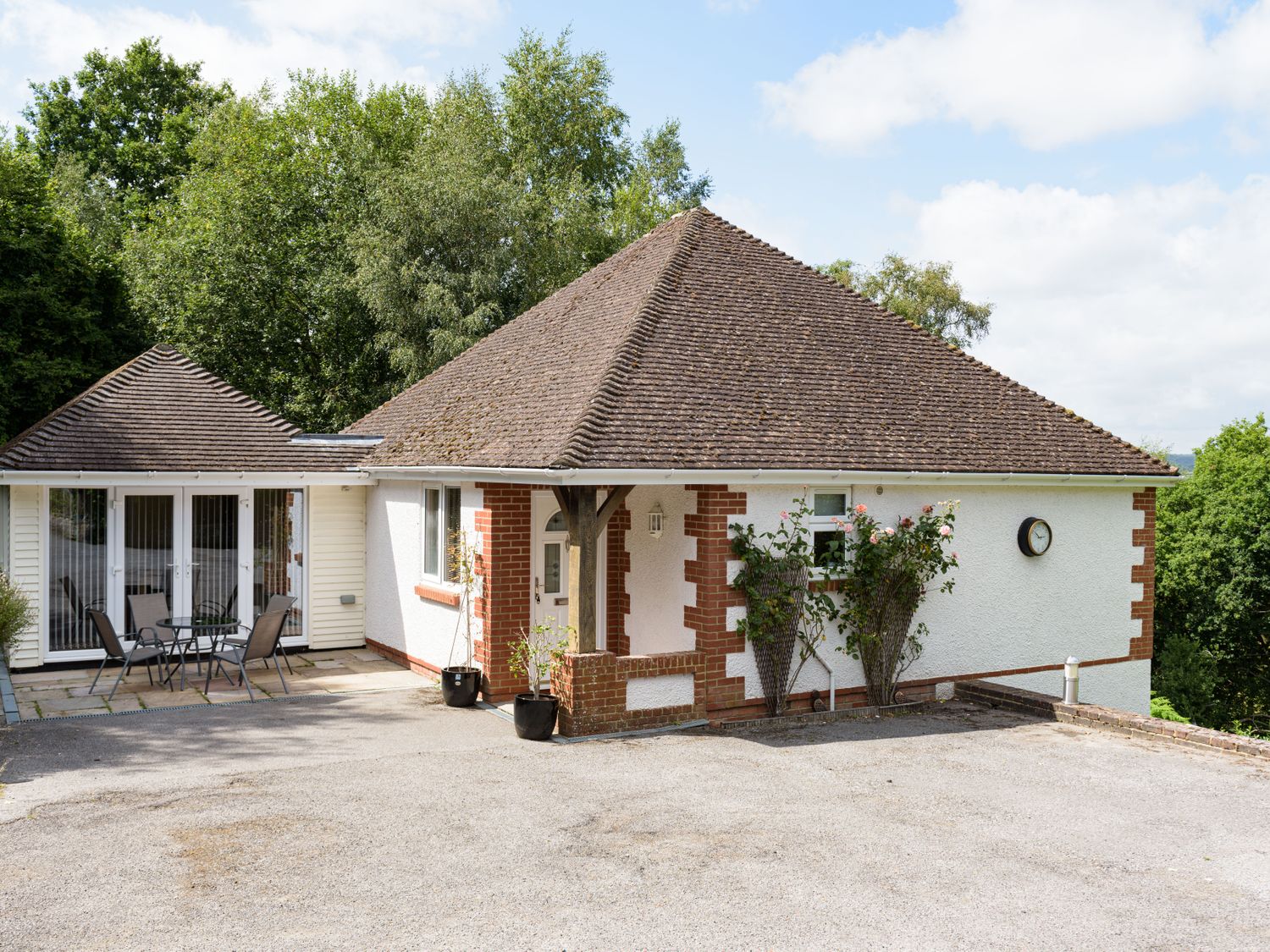 A single story house with a tiled roof and white walls with red brick accents outside patio furniture and green trees at Heath Retreat in Buxted