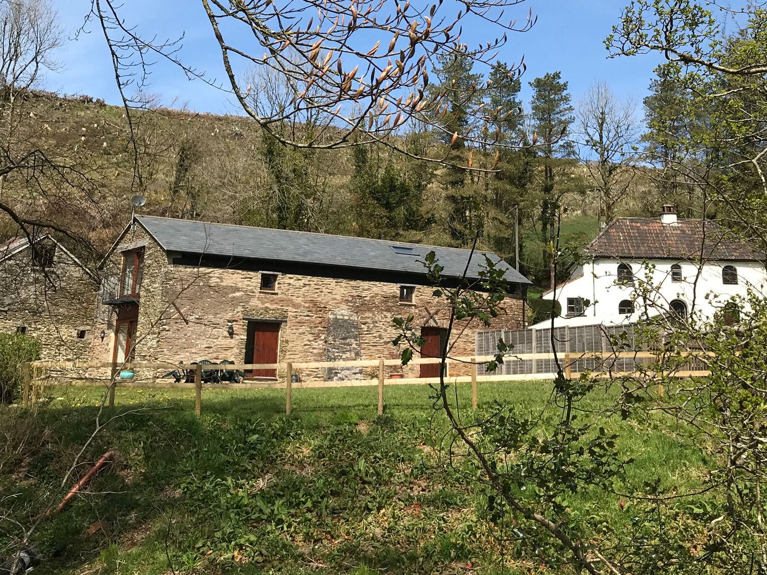 A stone building and a white house on grassy land with trees and a hill in the background at Red Deer Cottage in Wheddon Cross