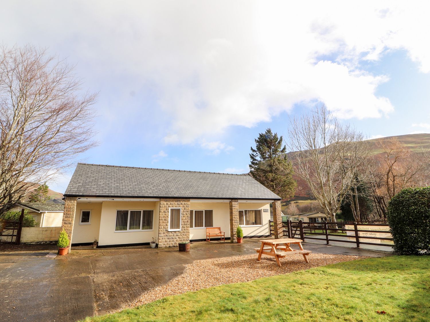 A single story building with outdoor bench and picnic table on a paved and gravel area with grass and trees surrounding at Awel Y Berwyn in Llangynog