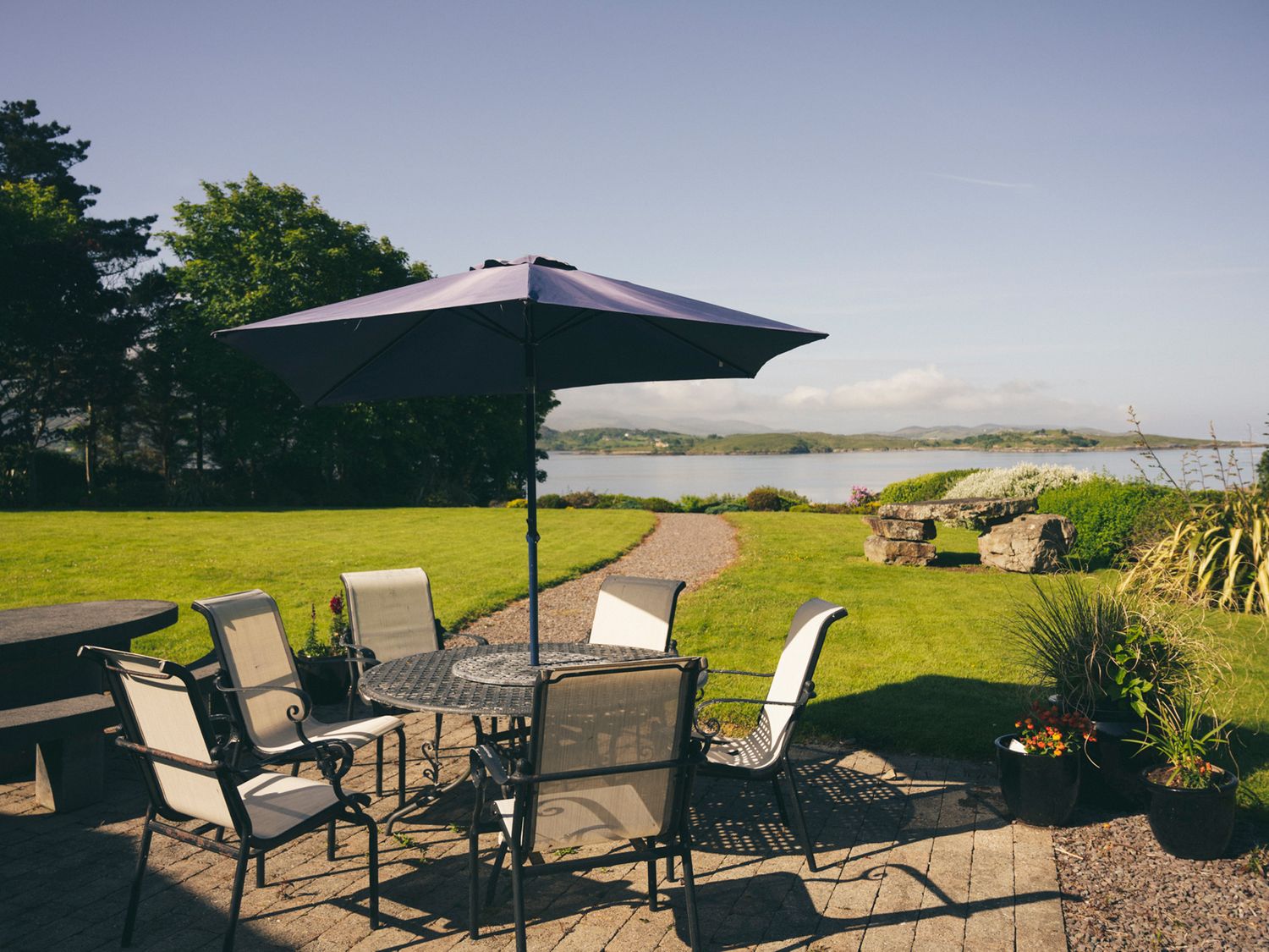 An outdoor patio with a round table and six chairs under a large umbrella overlooking a lawn and a body of water at Inbear Sceine Laughaunacreen near Tuosist County Kerry