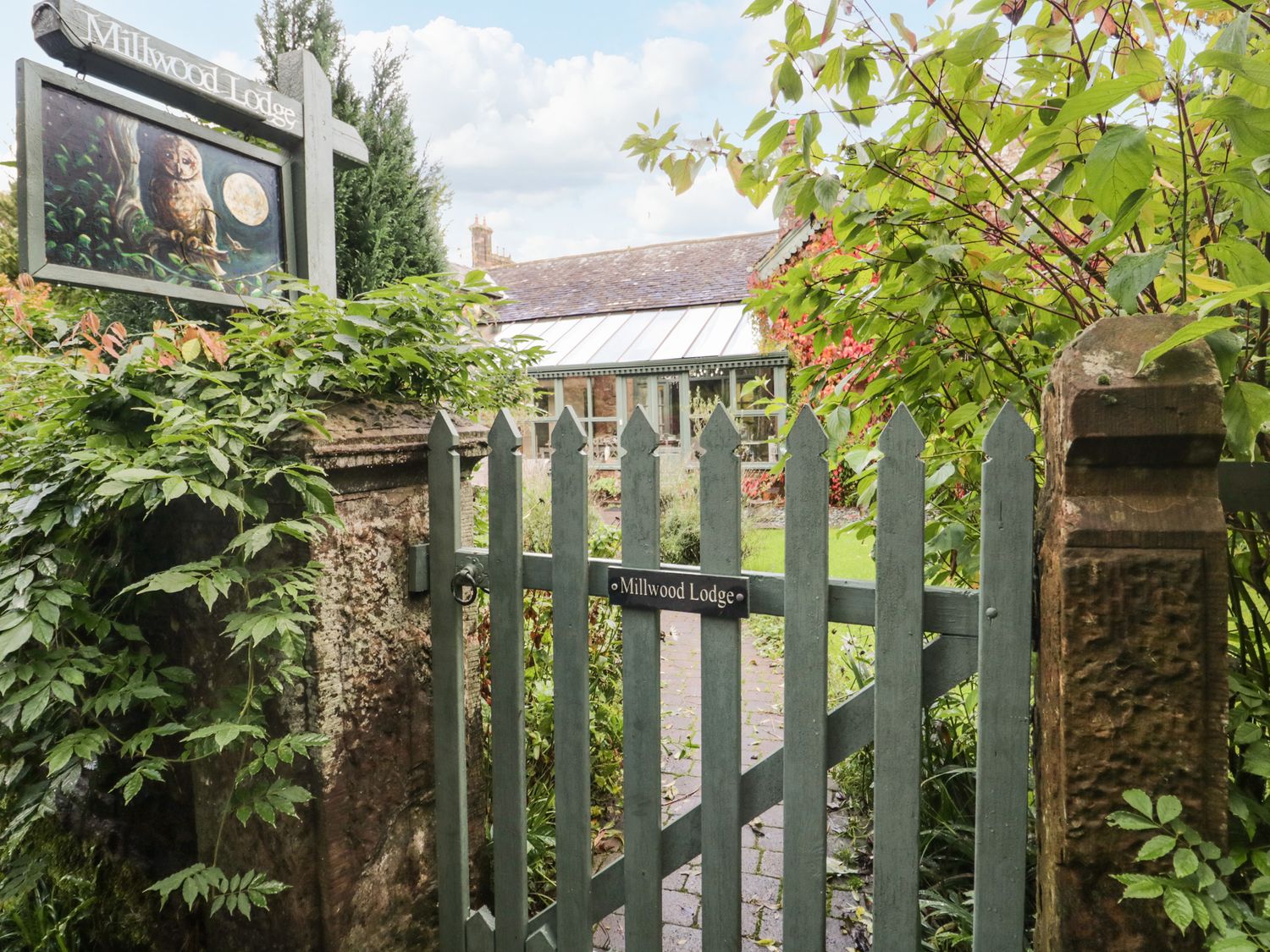 A wooden gate with a sign reading Millwood Lodge surrounded by plants and a building in the background at Millwood Lodge in Barrow In Furness