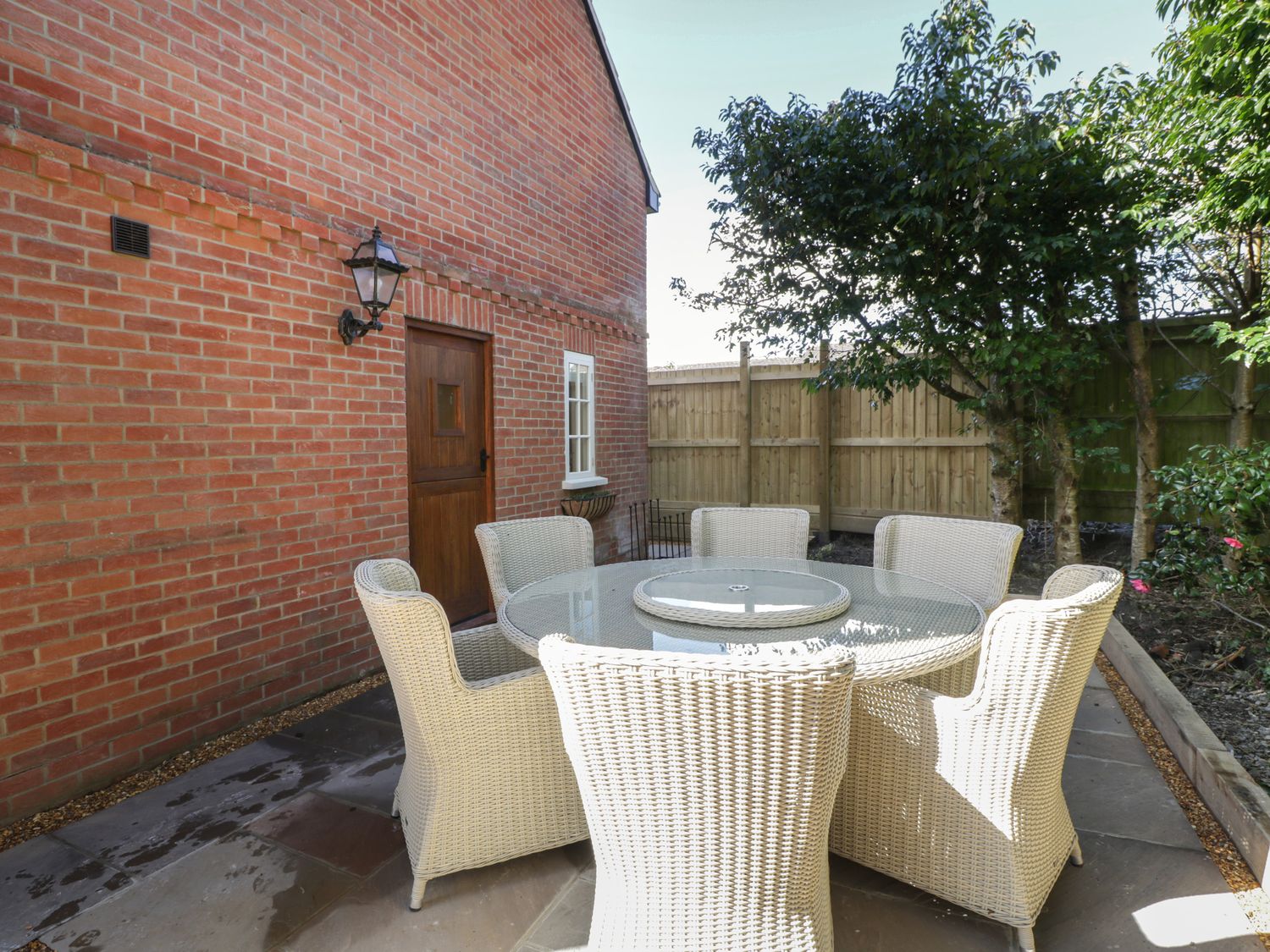 An outdoor patio with a round glass table and six wicker chairs near a brick wall and wooden door at The Coach House@15 in Warminster