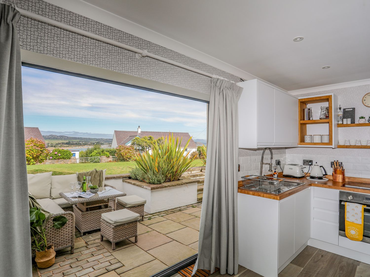A kitchen with white cabinets and wooden countertops opening to an outdoor seating area with a table and cushioned chairs at Trem Y Borth Apartment in Bwlchtocyn near Abersoch