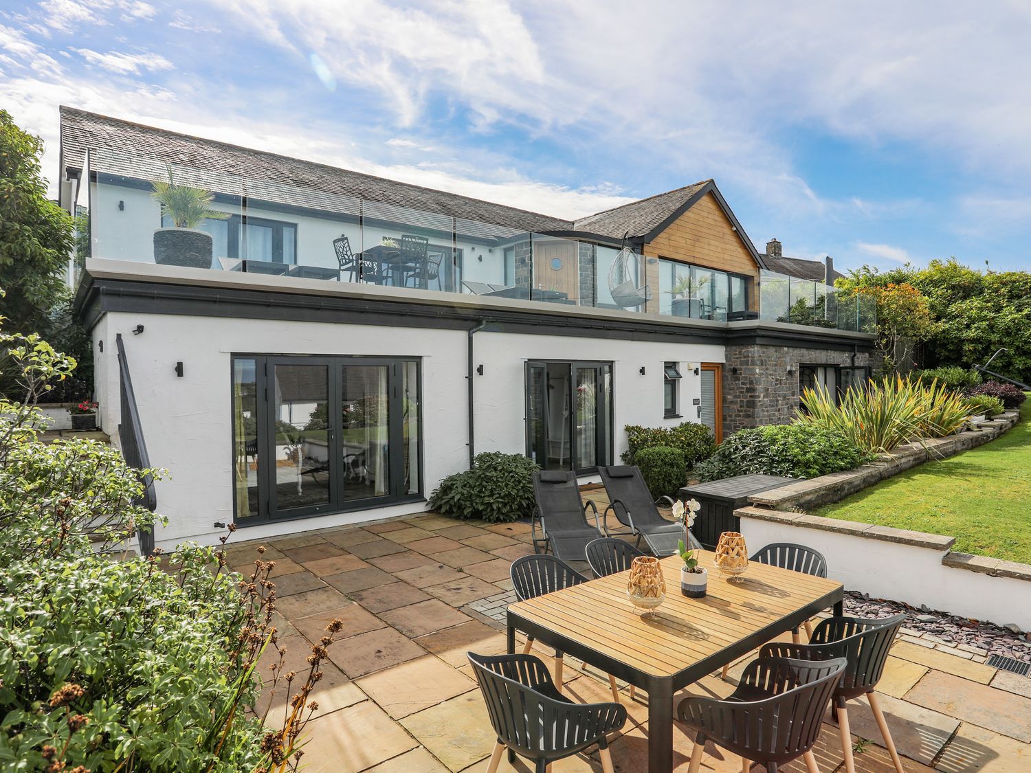 A patio area with a wooden dining table and chairs and reclining chairs outside a two-story house with glass balcony railings at Trem Y Borth Apartment in Bwlchtocyn near Abersoch