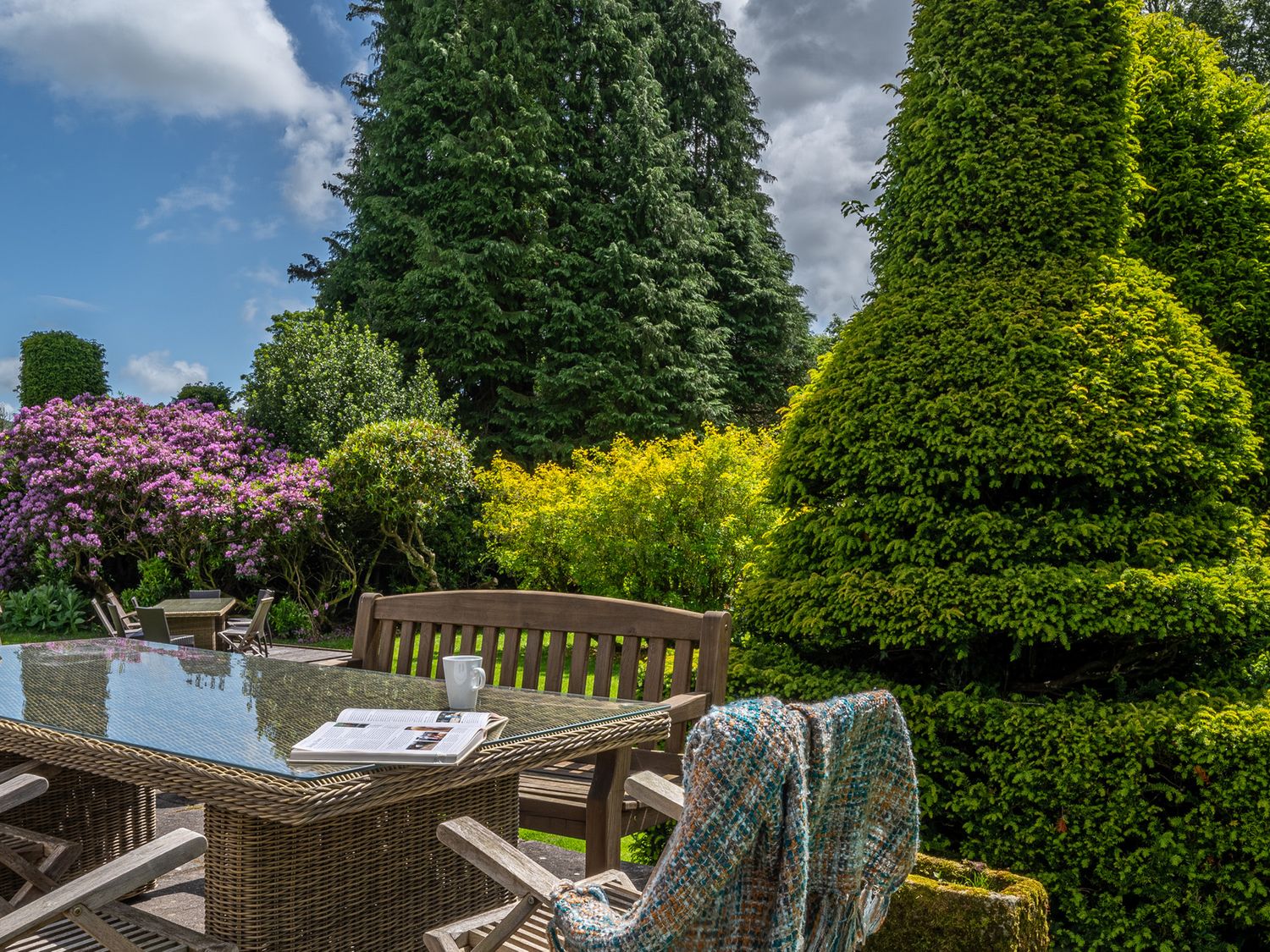 An outdoor garden with a glass table and wooden chairs surrounded by trees and bushes at Low Graythwaite Hall in Hawkshead