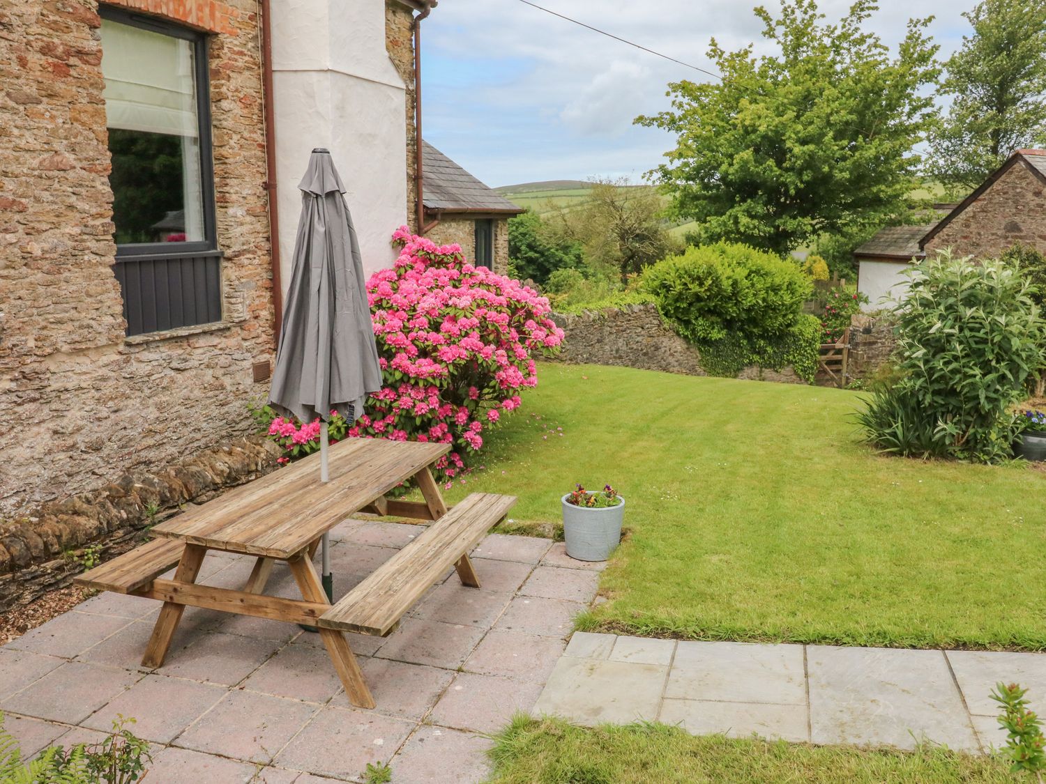 A garden area with wooden picnic table and closed umbrella by a stone house with plants and lawn at Ash Cottage in Combe Martin