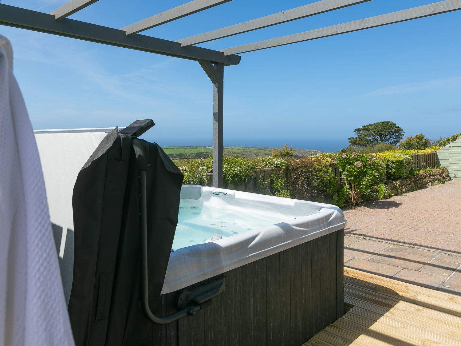 An outdoor hot tub under a wooden pergola with a garden and distant sea view at Broadlands Bosavern near St Just