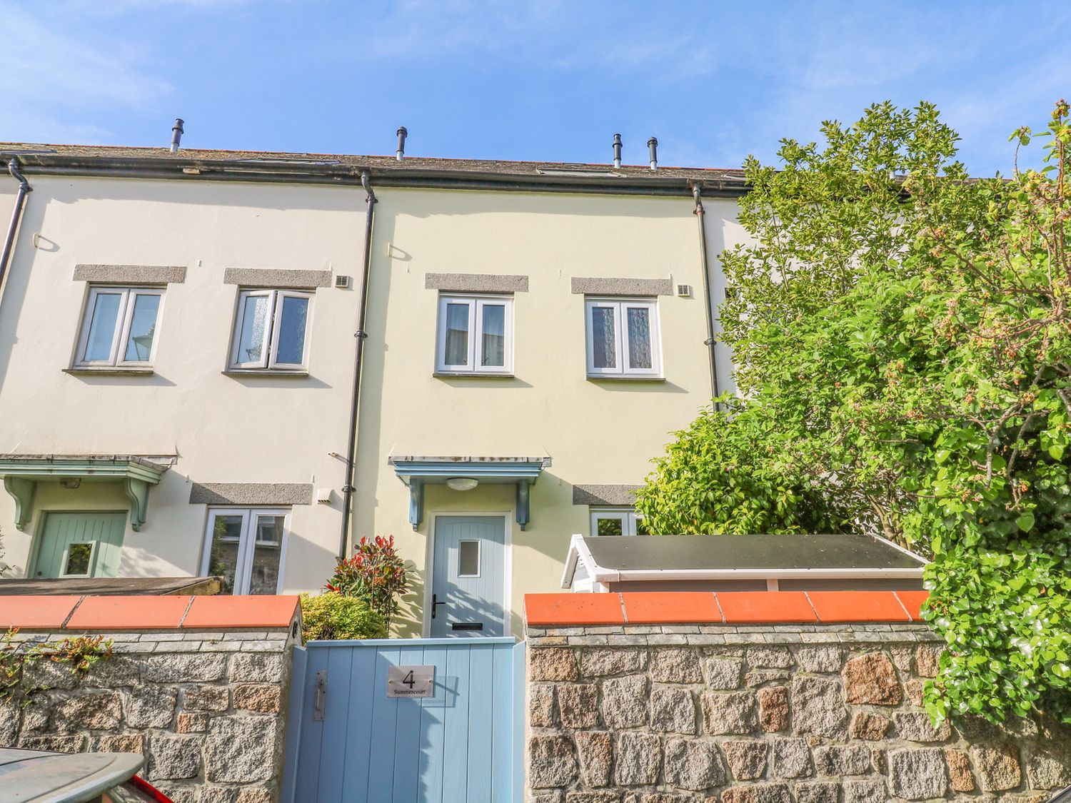 The front view of a two-story terraced house with a blue gate and door and a stone wall with plants at Riverside Summercourt in Penryn