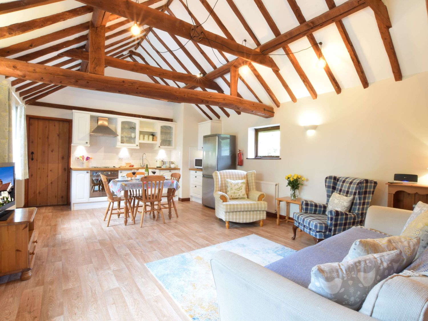 A living area with wooden beams on the ceiling a kitchen with dining table and chairs an armchair and a sofa with cushions at The Stables in Polstead