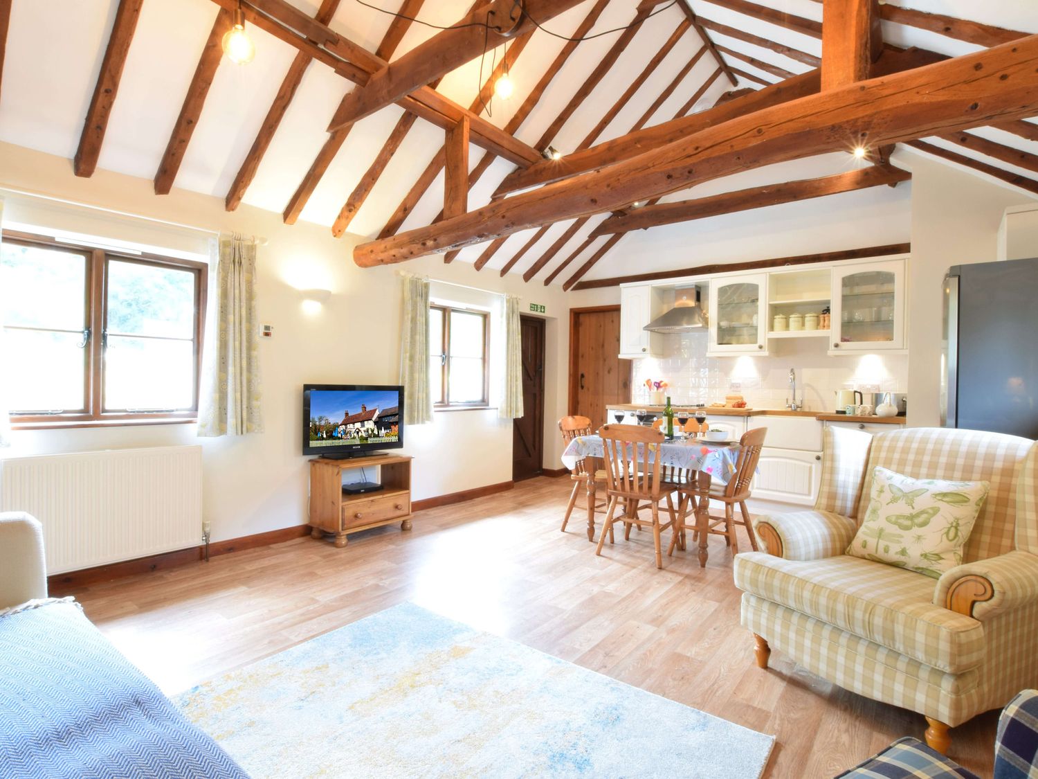 An open plan living area with wooden beams dining table chairs kitchen cabinets television armchair and rug at The Stables in Polstead