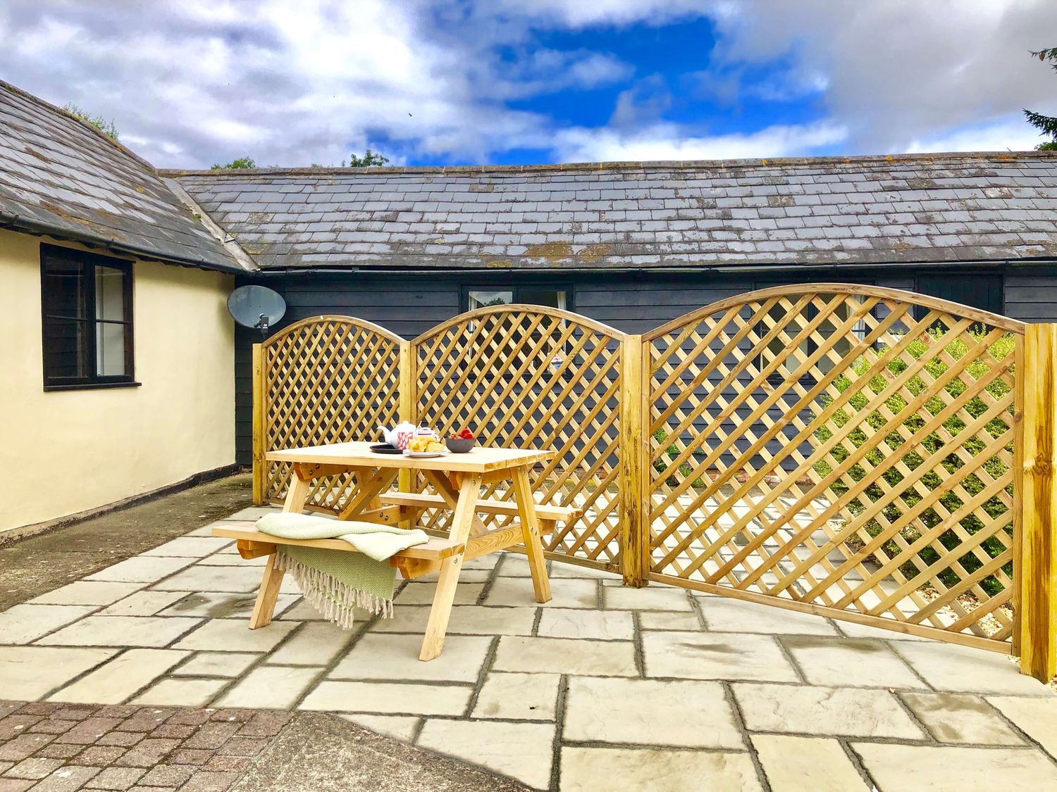 A wooden picnic table with food and a blanket on the bench on a stone patio with lattice fencing and buildings in the background at The Stables in Polstead