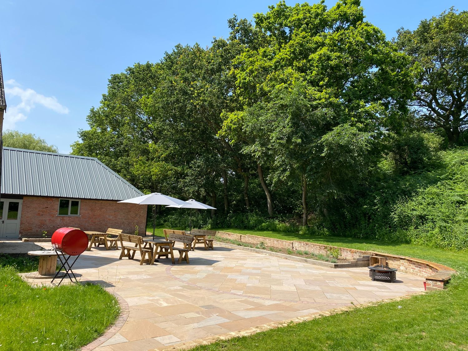 An outdoor patio area with wooden benches and tables under white umbrellas next to a brick building with a metal roof surrounded by grass and trees at The Stables in Polstead