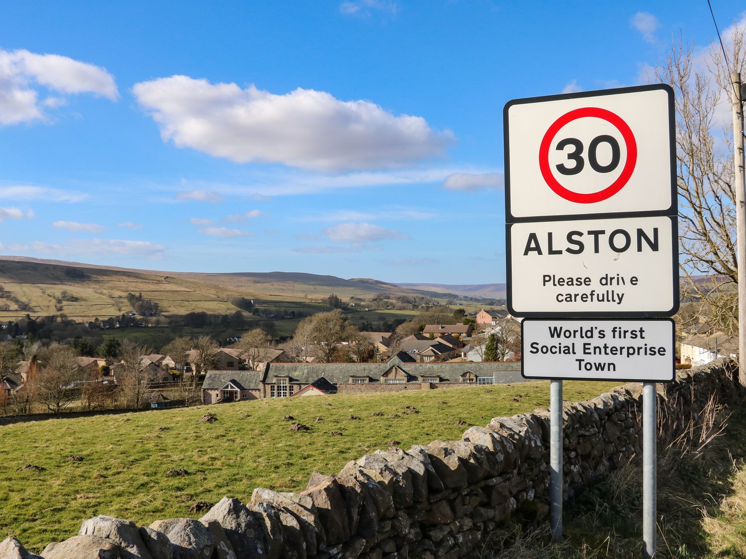 A speed limit sign and town sign for Alston in a rural area with houses and hills in the background at Cross View Cottage in Alston