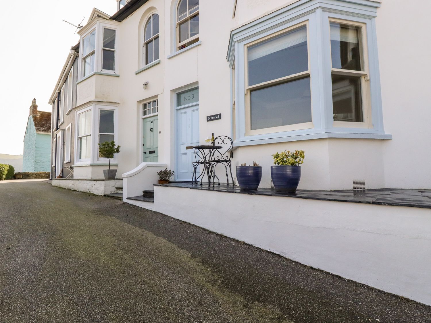 A terrace with a small table and two chairs outside a white house with blue window frames at Driftwood in Fowey