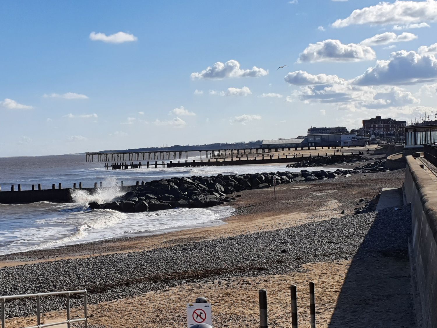 A beach with rocky shore and waves hitting rocks with a pier and buildings in the background at Seaside Retreat in Lowestoft