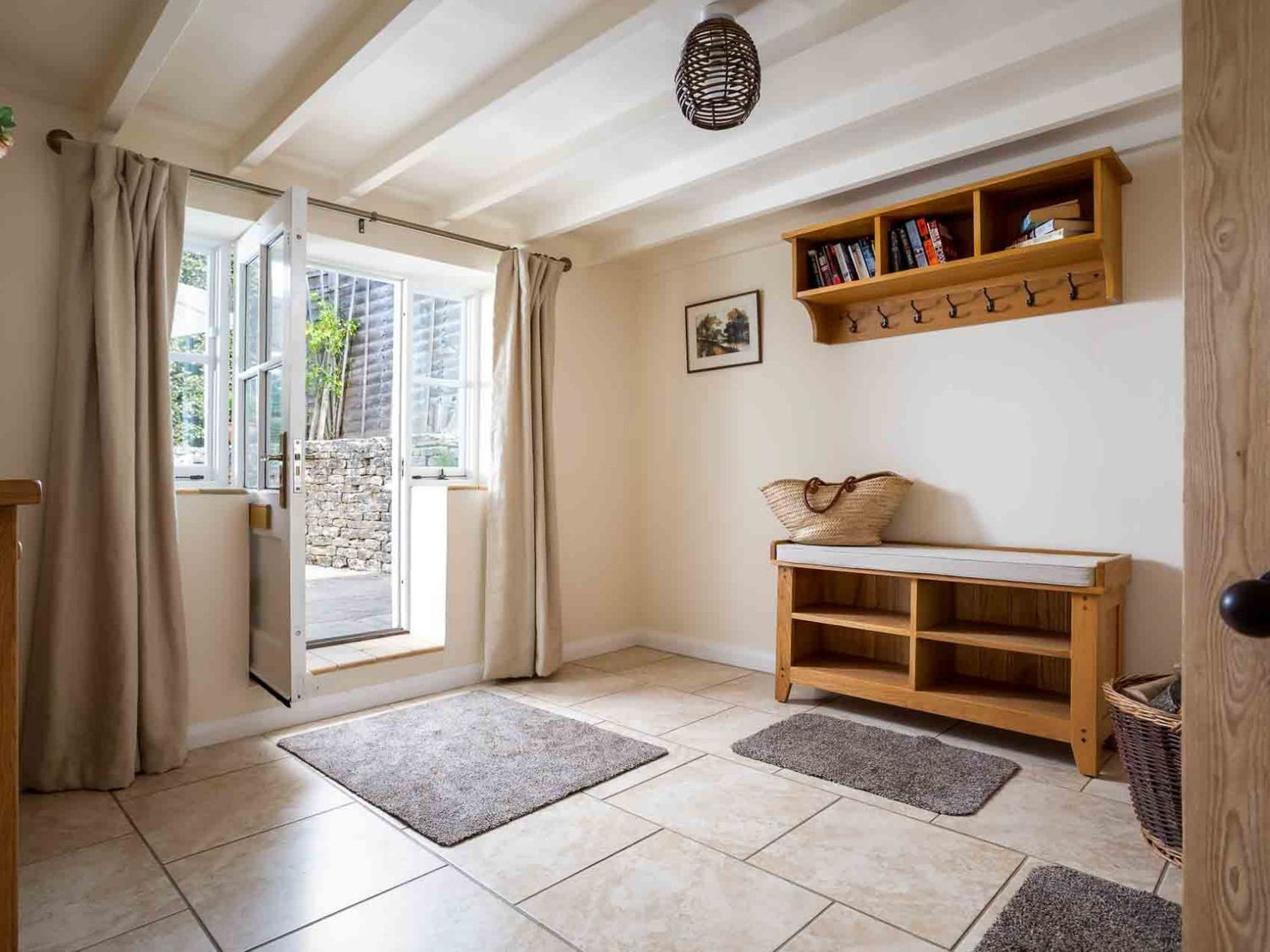 A tiled mudroom with an open door with curtains mats a wooden bench with storage and a wall-mounted wooden shelf with hooks and books at Weir Cottage in Bourton-on-the-Water