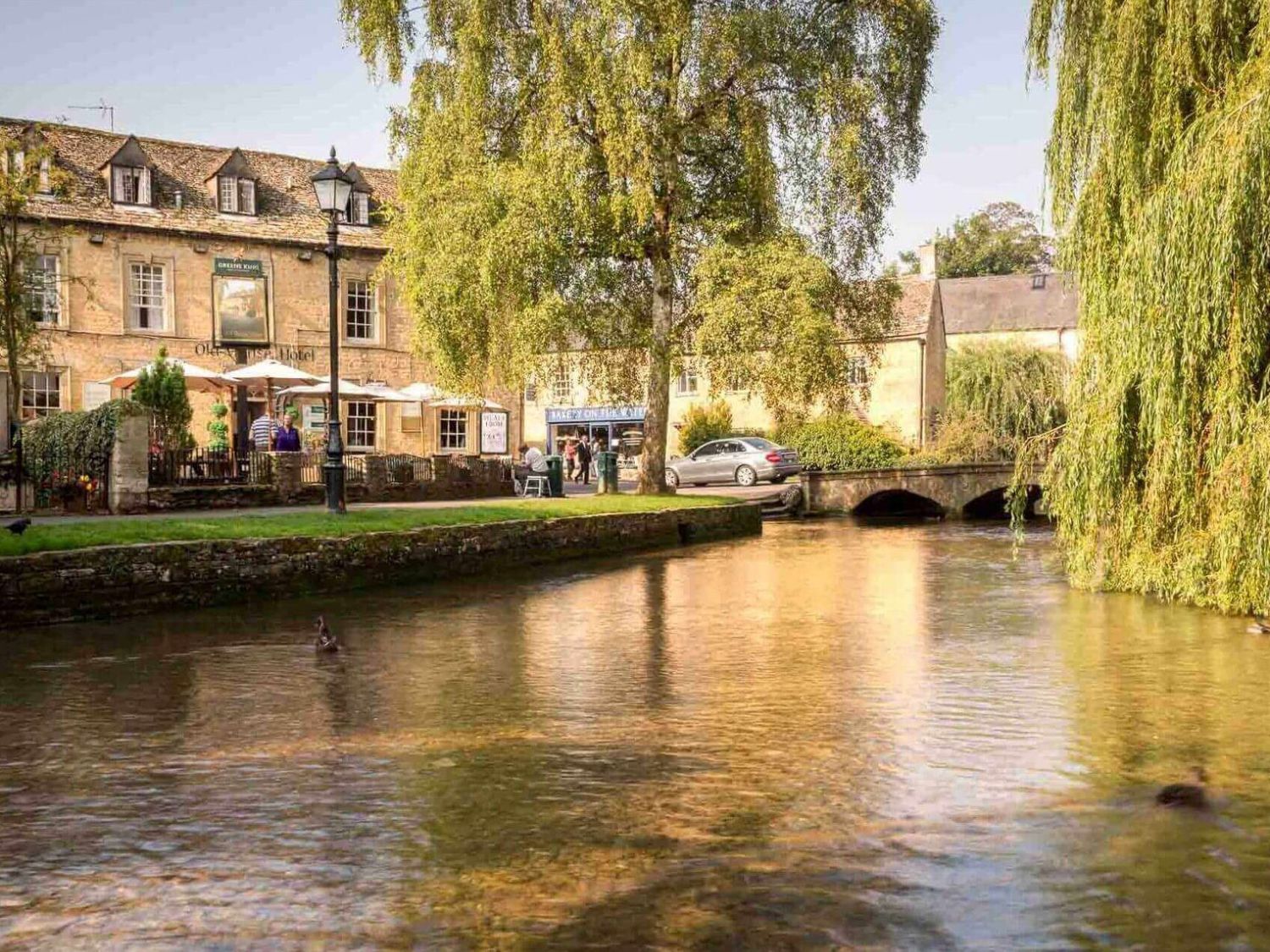A river flowing beside a stone building with umbrellas and a car on a small bridge at Weir Cottage in Bourton-on-the-Water