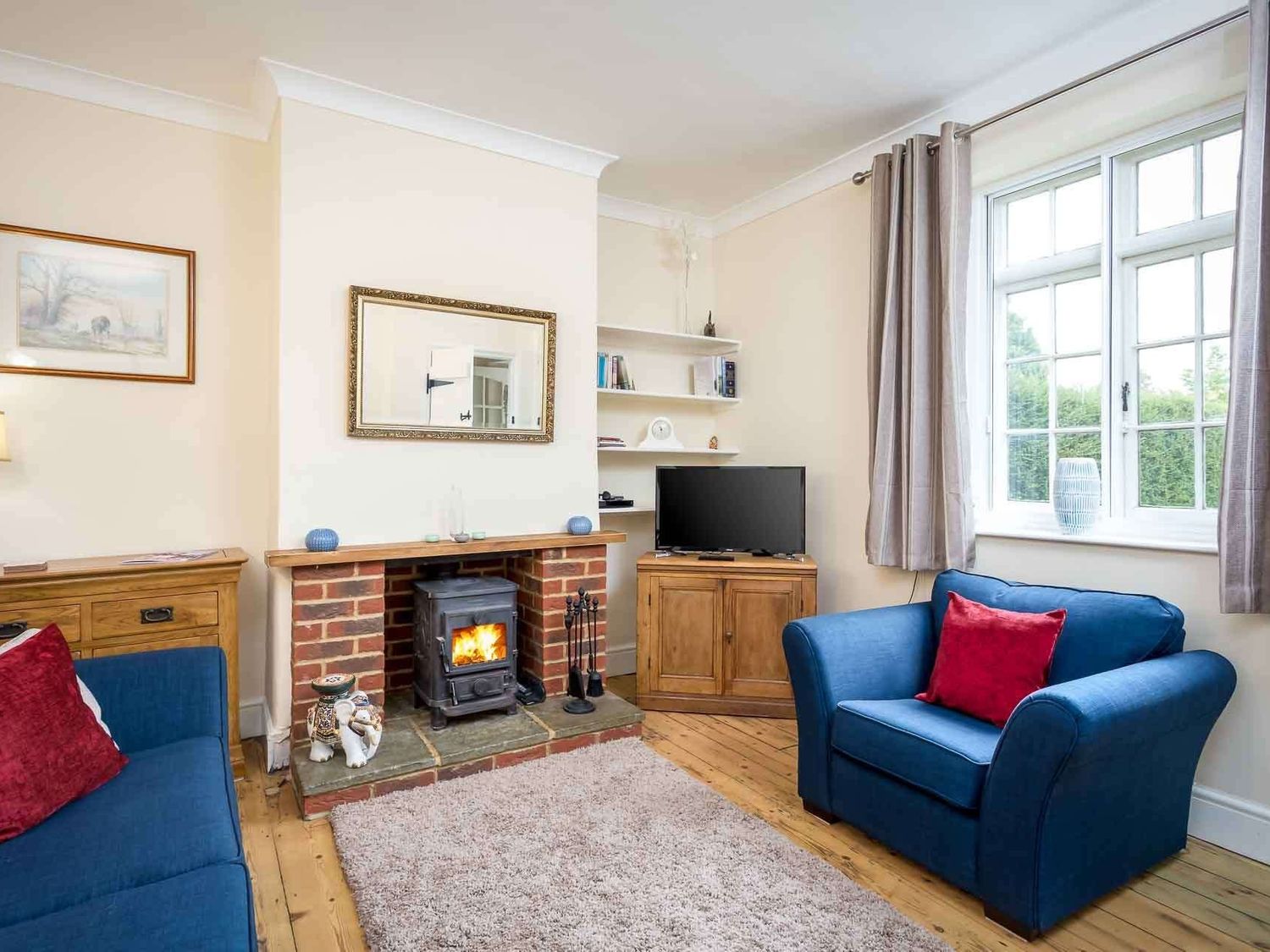 A living room with blue armchairs red cushions a fireplace and a television at Swan View in Southrop