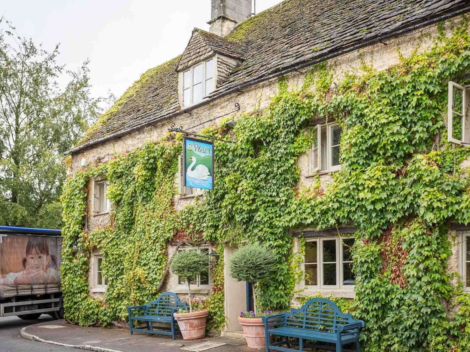A stone building covered in ivy with blue benches and potted plants outside at Swan View in Southrop
