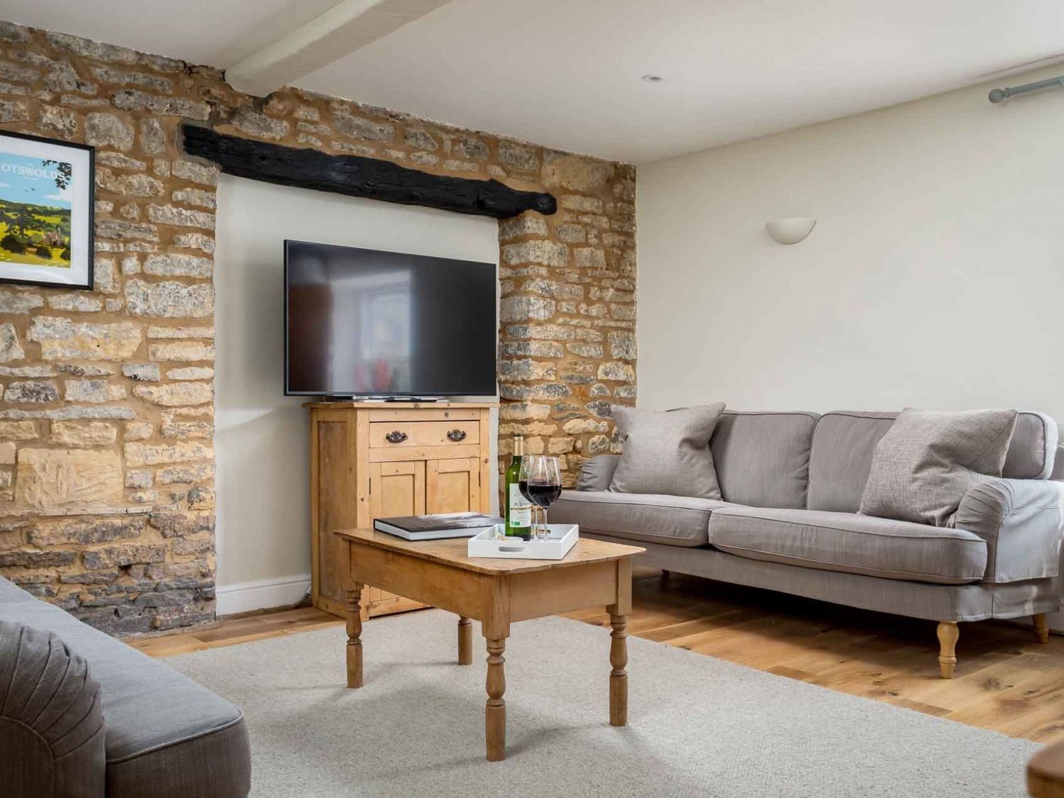 A living room with two grey sofas a wooden coffee table with a book and wine glasses a wooden cabinet with a TV on top and a stone feature wall at High Ridge in Milton-under-Wychwood