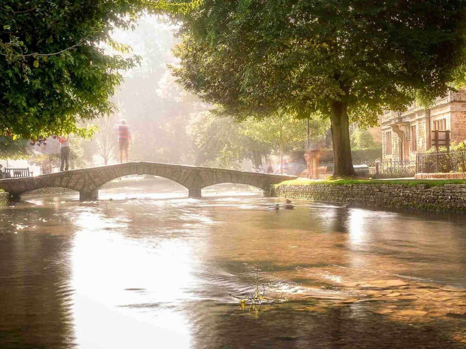 A stone arch bridge over a river with trees and a building along the riverbank at High Ridge in Milton-under-Wychwood