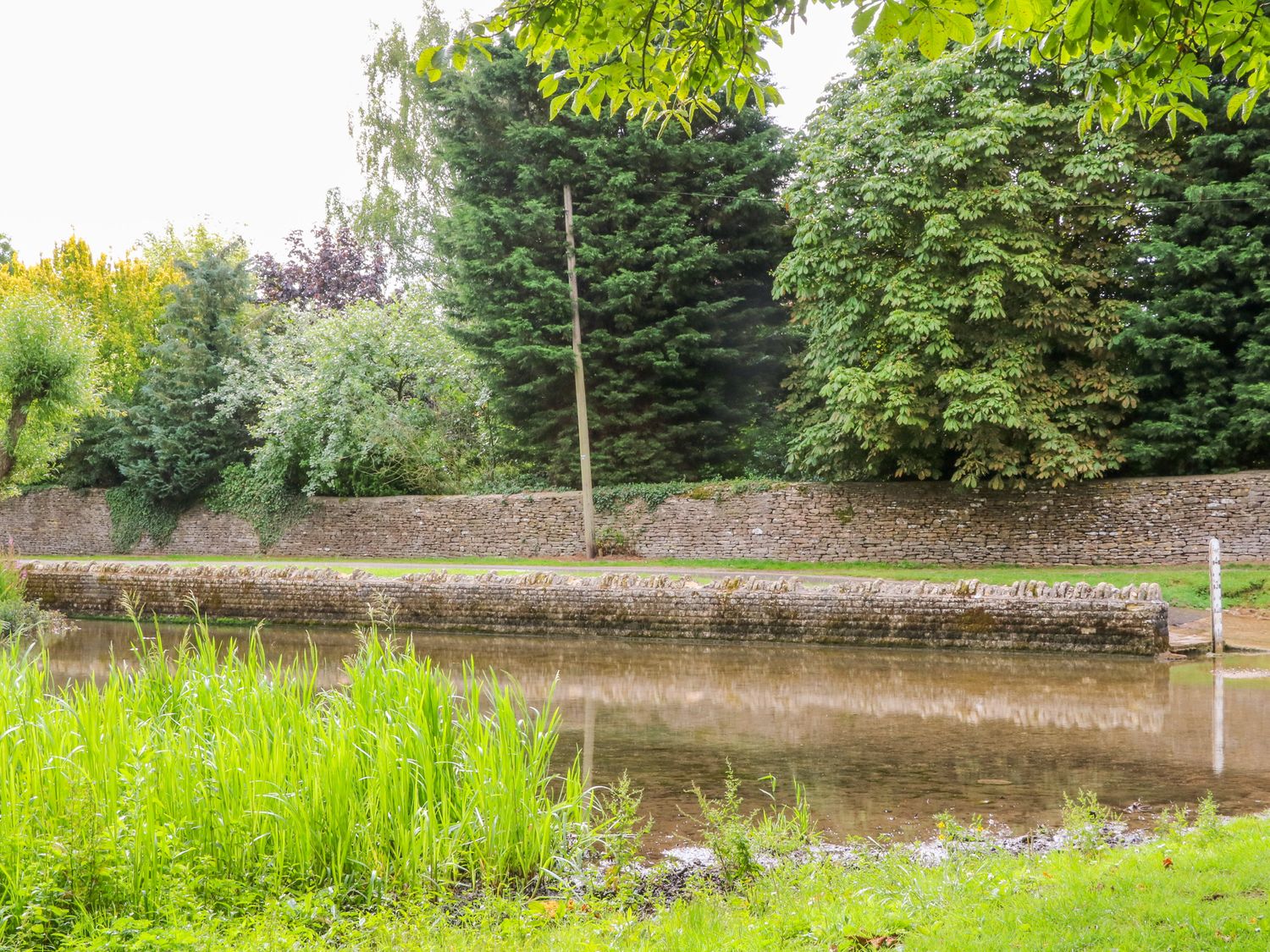 A stone wall along a narrow body of water with tall grass and trees at Rood Cottage in Shilton near Burford