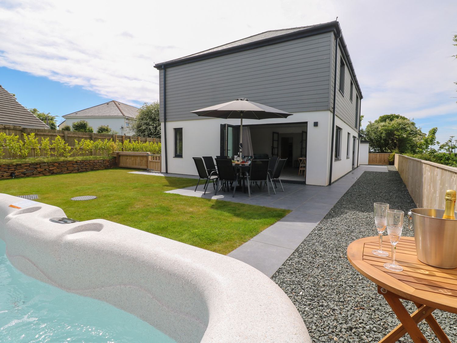 A backyard with a hot tub a patio table and chairs under an umbrella and a wooden table with glasses and a champagne bucket at Tredarn in Connor Downs