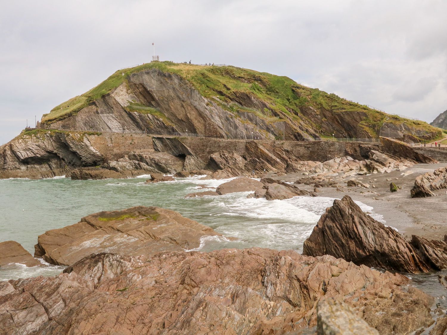 A rocky coastline with waves hitting the shore and a green hill in the background at 65 Channel View in Ilfracombe